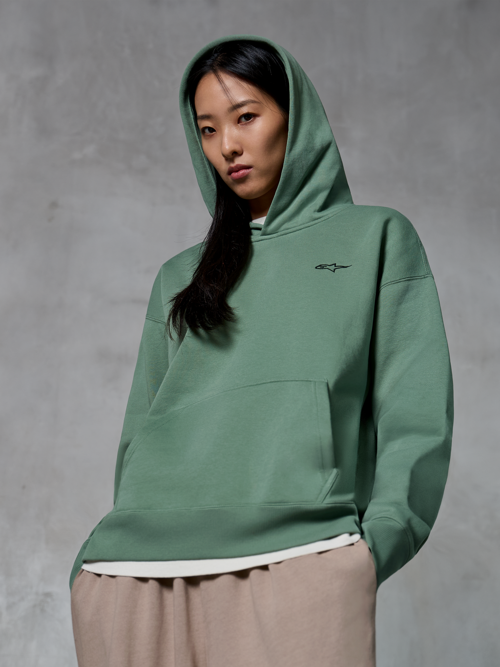 Sweat à capuche Alpinestars Essentials Ageless, streetwear décontracté, vert Cactus avec un petit logo noir, porté avec la capuche relevée, poche kangourou à l'avant, finition délavée, associé à un pantalon beige clair