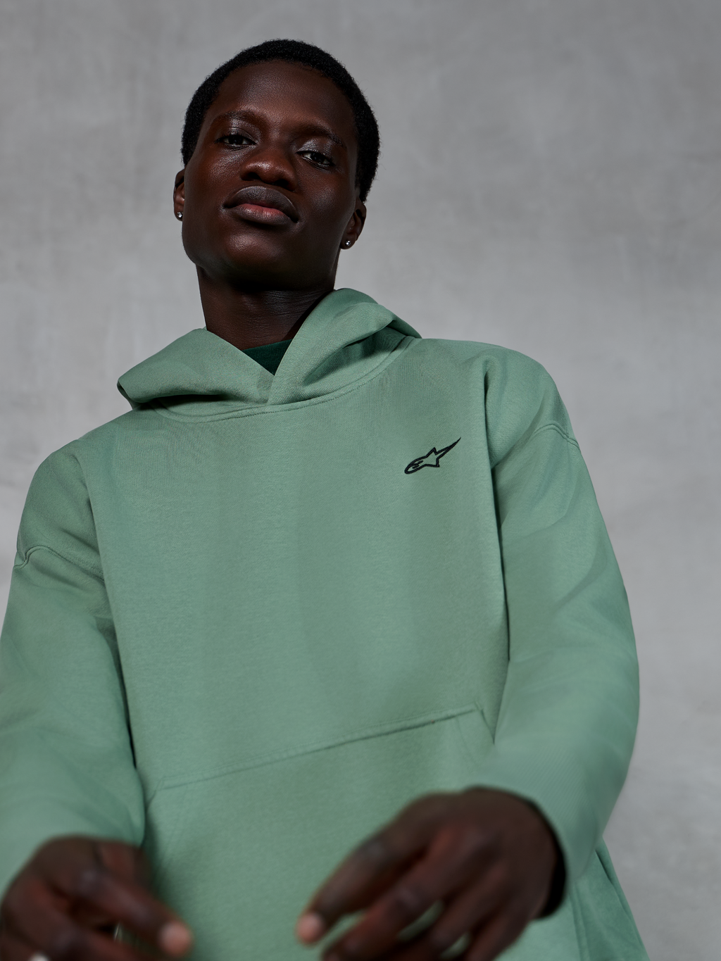Sweat à capuche Alpinestars Essentials Ageless, vêtement de sport décontracté de couleur vert Cactus, porté par un mannequin, avec un petit logo Alpinestars noir sur la poitrine, molleton de haute qualité, coupe décontractée et esthétique épurée.