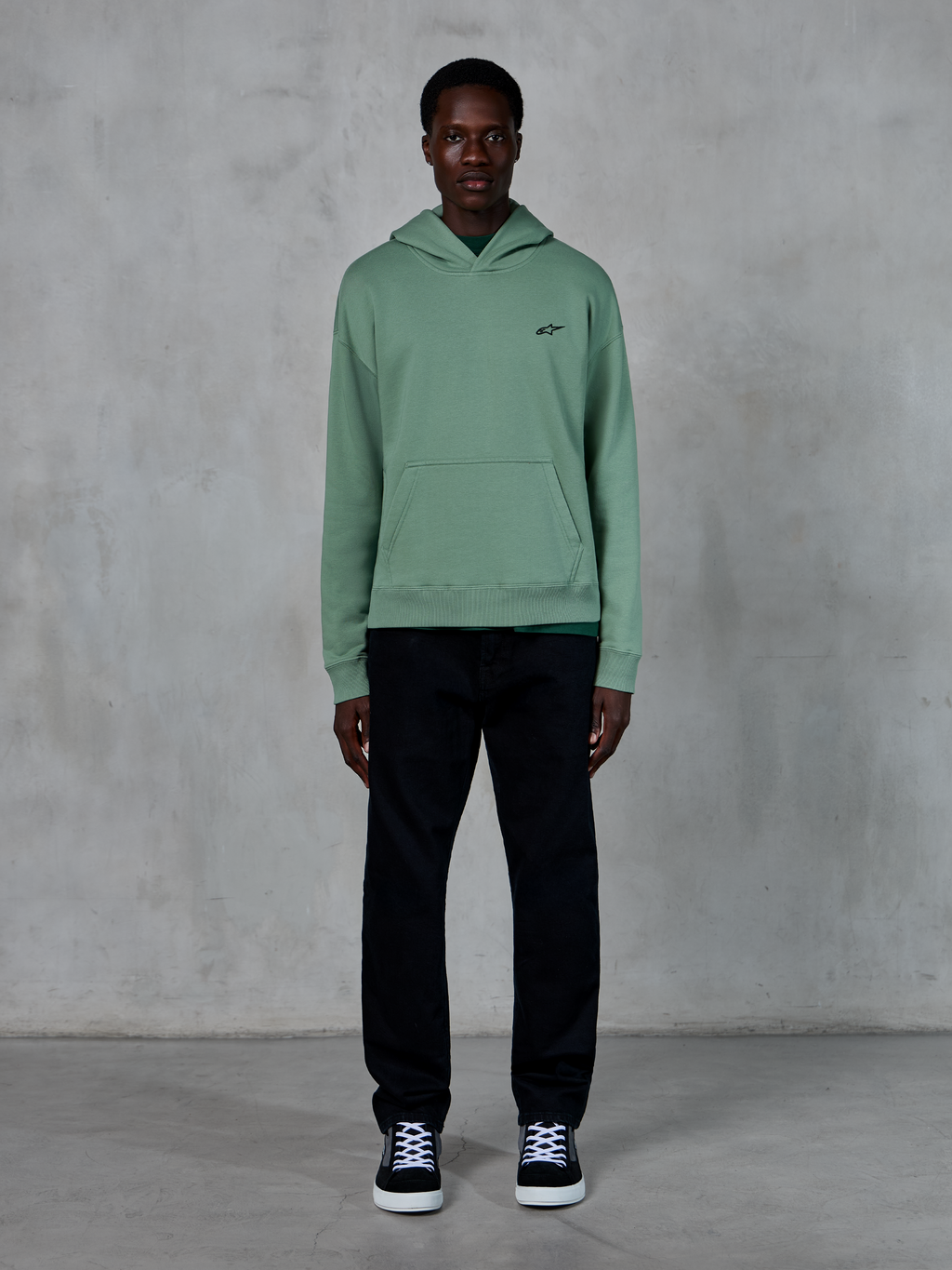 Sweat à capuche Alpinestars Essentials Ageless, vert Cactus, petit logo noir sur la poitrine, poche kangourou, style streetwear décontracté porté avec un pantalon noir et des baskets noires et blanches devant un mur texturé