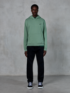 Sweat à capuche Alpinestars Essentials Ageless, vert Cactus, petit logo noir sur la poitrine, poche kangourou, style streetwear décontracté porté avec un pantalon noir et des baskets noires et blanches devant un mur texturé
