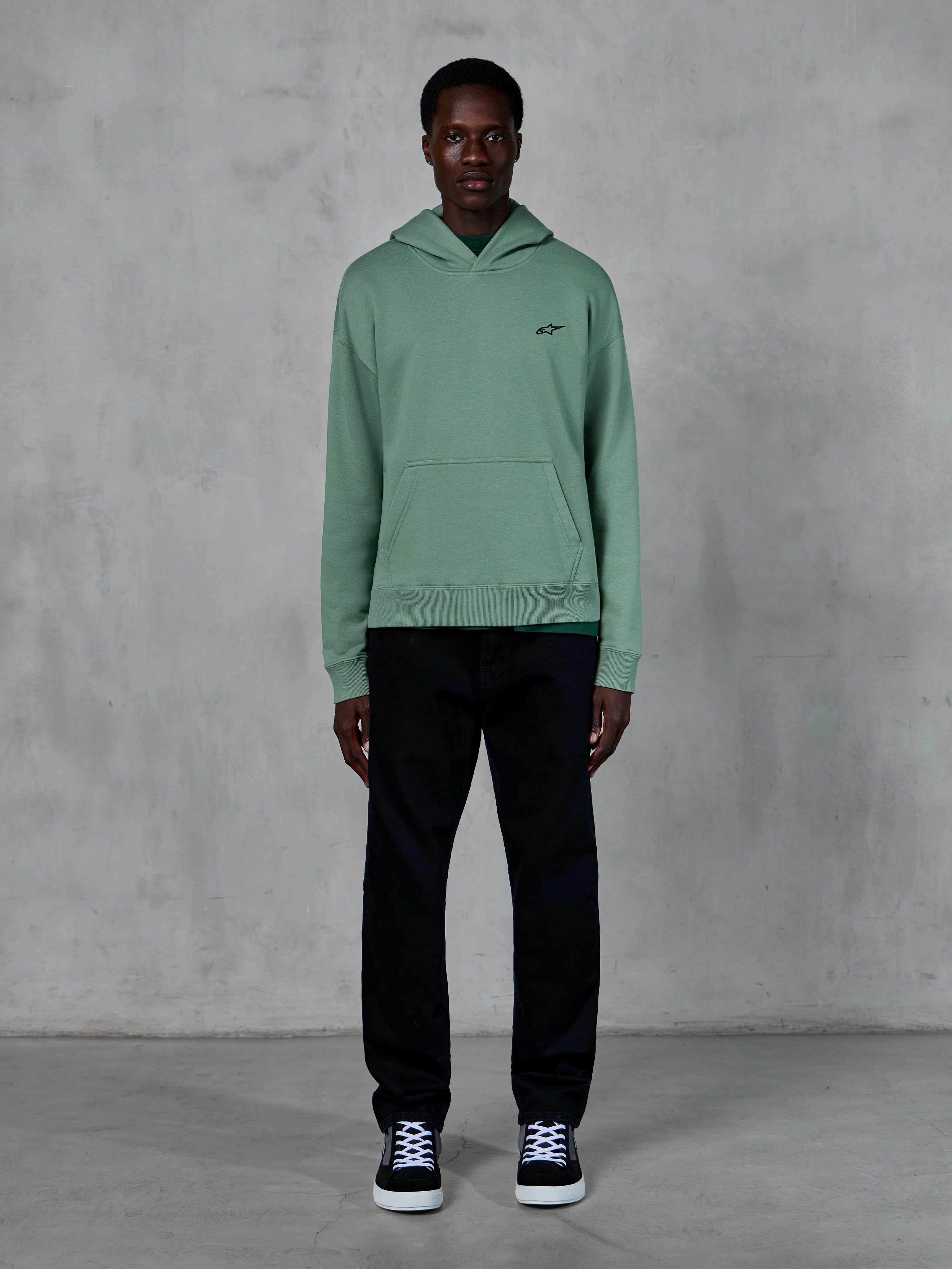 Sweat à capuche Alpinestars Essentials Ageless, vert Cactus, petit logo noir sur la poitrine, poche kangourou, style streetwear décontracté porté avec un pantalon noir et des baskets noires et blanches devant un mur texturé