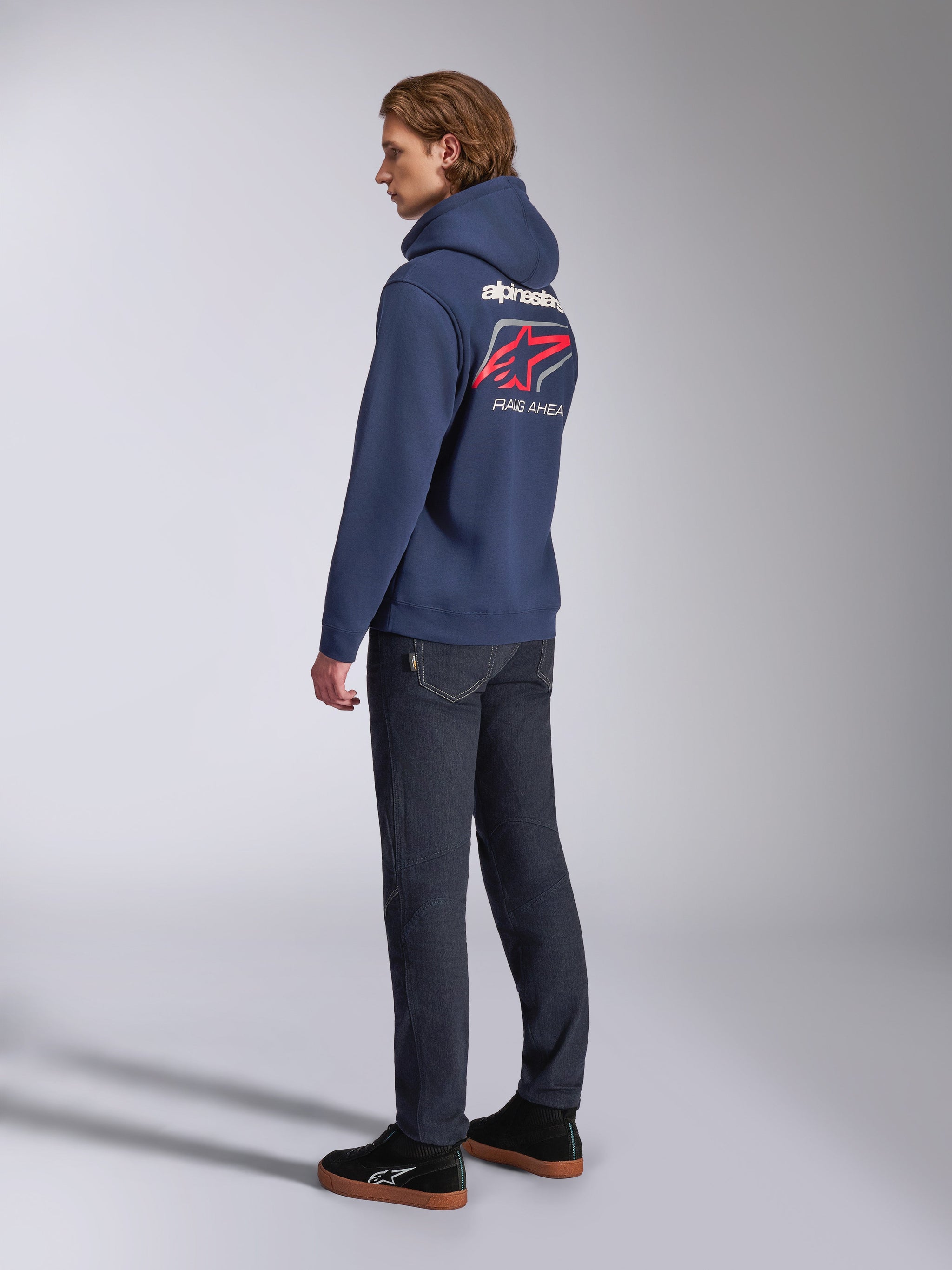 Alpinestars Formulation Hoodie, navy blue, back view showing white Alpinestars wordmark and red Astars logo with 'RACING AHEAD' text, worn with dark jeans and black sneakers, casual motorsport-inspired lifestyle apparel