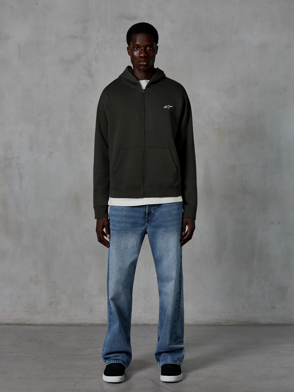 Sweat à capuche zippé Alpinestars Essentials Ageless, hoodie entièrement zippé, noir, avec un petit logo Alpinestars blanc sur la poitrine, porté par un mannequin avec un t-shirt blanc et un jean bleu, coupe décontractée, style streetwear décontracté motorsport