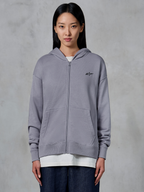 Sweat à capuche zippé Alpinestars Essentials Ageless, Silver, streetwear décontracté, sweat en polaire à fermeture éclair intégrale avec petit logo Alpinestars noir sur la poitrine, poche kangourou divisée, porté par un mannequin sur un t-shirt blanc et un jean foncé