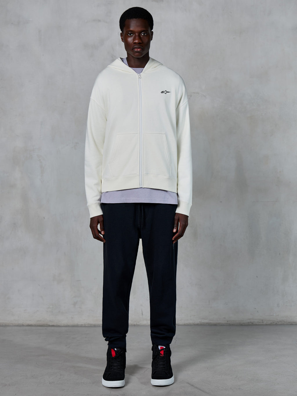 Alpinestars Essentials Ageless Zip Hoodie, sweat à capuche zippé blanc cassé, coupe décontractée avec petit logo noir sur la poitrine, porté avec un pantalon de jogging noir et des baskets, style streetwear décontracté inspiré du sport automobile