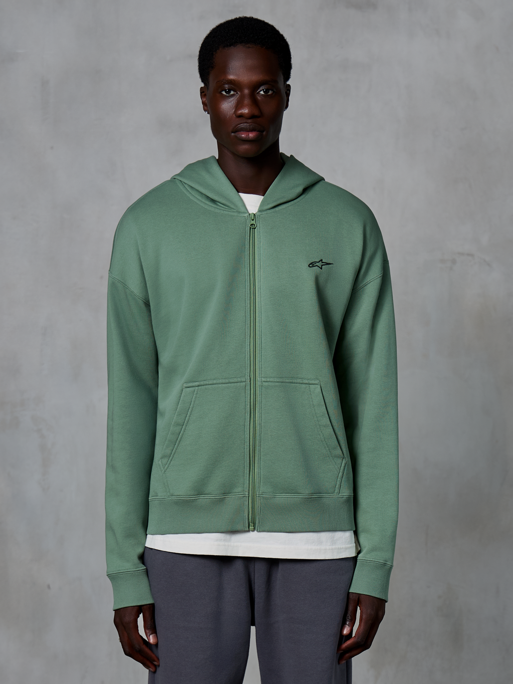 Sweat à capuche zippé Alpinestars Essentials Ageless, vêtement streetwear décontracté, vert Cactus, avec un logo brodé discret sur la poitrine, des poches kangourou divisées et des poignets côtelés, porté par un mannequin homme sur un fond gris texturé