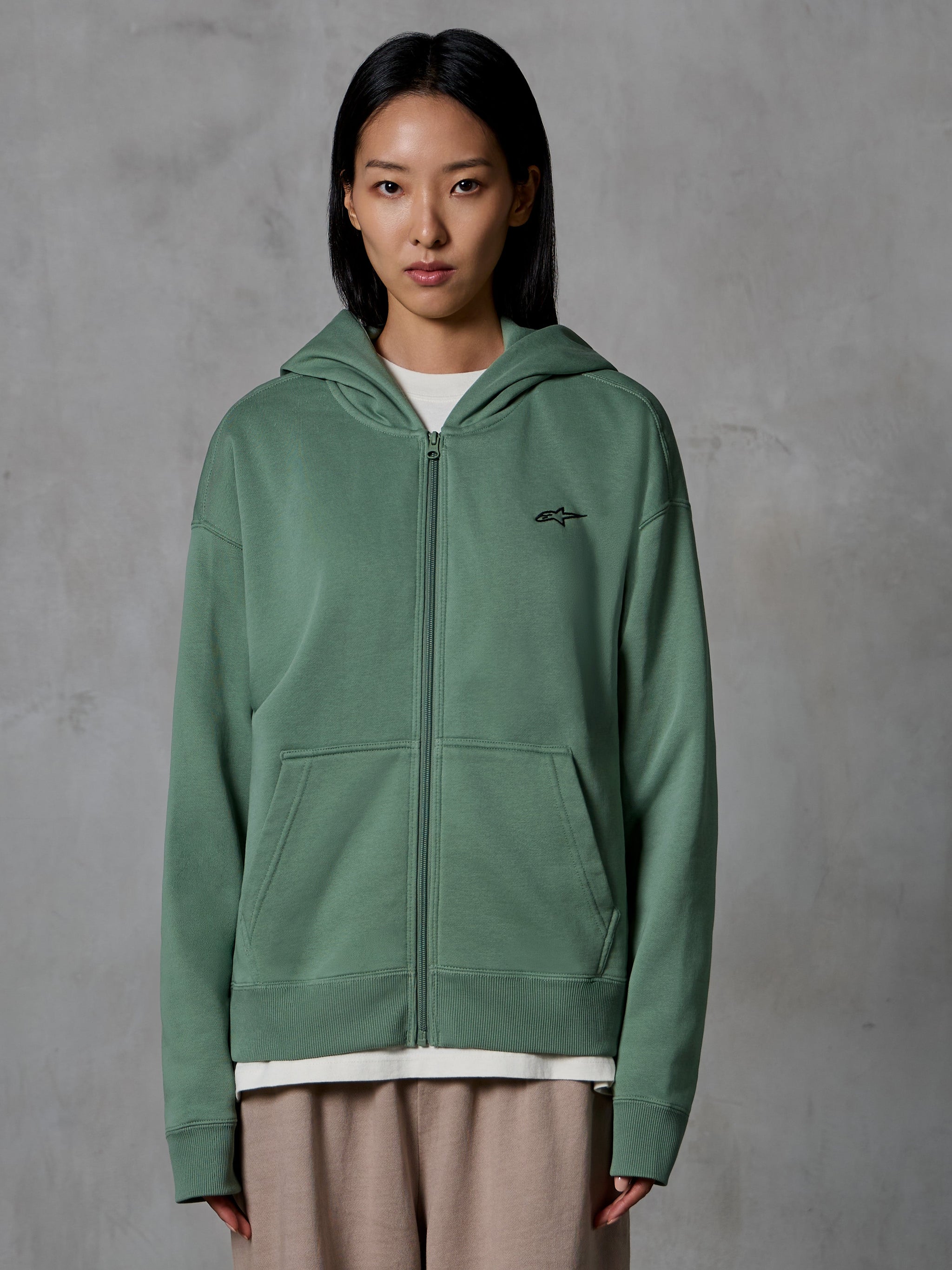 Sweat à capuche zippé Alpinestars Essentials Ageless, vert cactus, streetwear décontracté porté par un mannequin femme, sweat en polaire zippé avec petit logo noir sur la poitrine, poches avant, coupe décontractée pour un usage quotidien