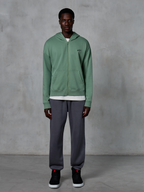 Sweat à capuche zippé Alpinestars Essentials Ageless, sweat à capuche zippé, vert cactus, porté par un mannequin avec un jogging gris foncé et des baskets noires, arborant un petit logo Alpinestars noir sur la poitrine, coupe décontractée, style streetwear décontracté