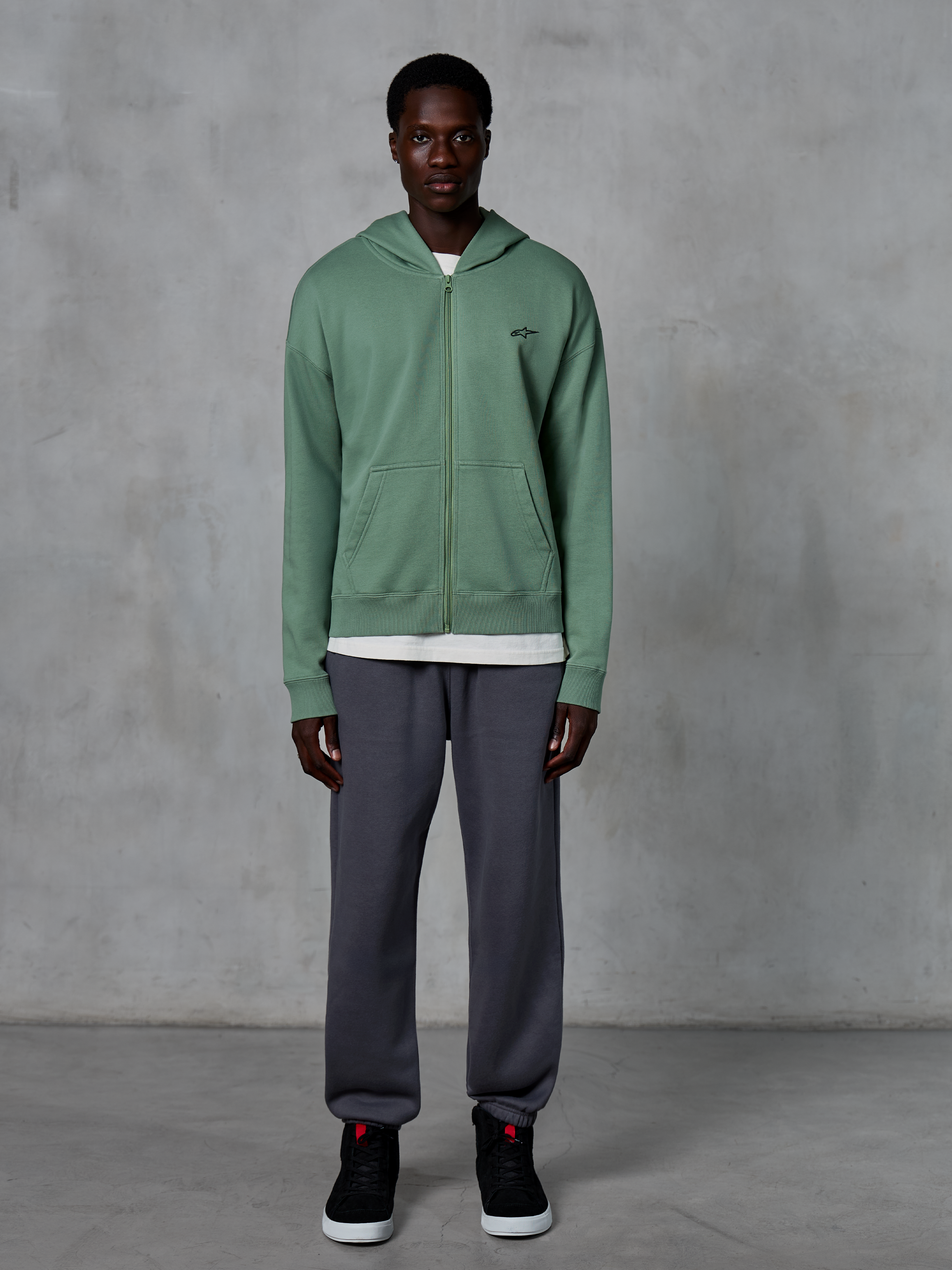 Sweat à capuche zippé Alpinestars Essentials Ageless, sweat à capuche zippé, vert cactus, porté par un mannequin avec un jogging gris foncé et des baskets noires, arborant un petit logo Alpinestars noir sur la poitrine, coupe décontractée, style streetwear décontracté
