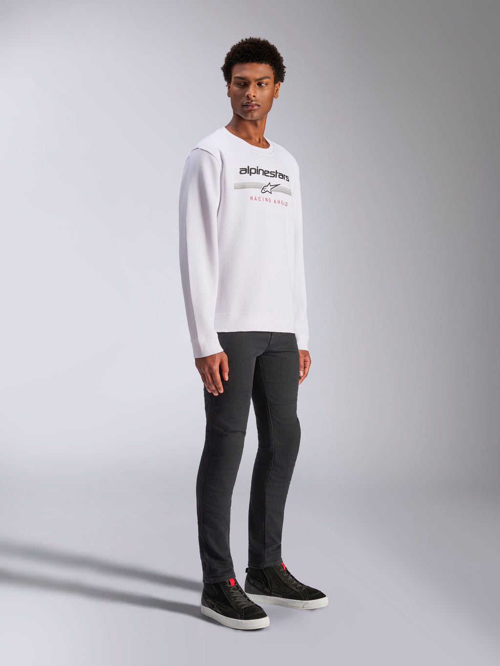 Alpinestars Bettern Crew, sweat-shirt blanc à col rond avec logo Alpinestars noir et texte rouge 'Racing Ahead', style streetwear décontracté inspiré du sport automobile, porté par un mannequin avec un pantalon sombre et des baskets noires