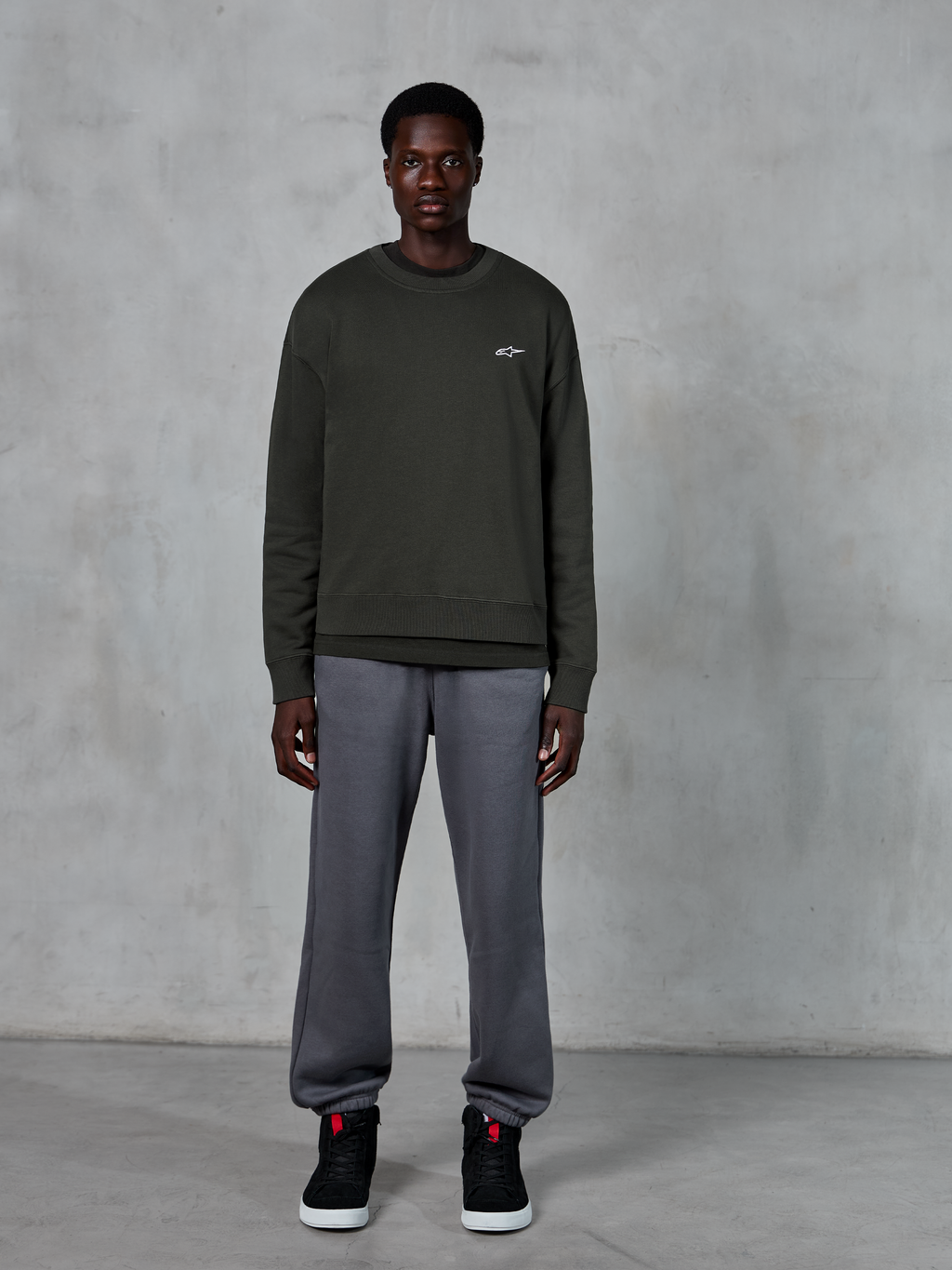 Alpinestars Essentials Ageless Crew, sweat-shirt à col rond vert foncé avec un petit logo blanc sur la poitrine, porté par un mannequin avec un pantalon de survêtement gris et des baskets montantes noires, vêtements lifestyle décontractés