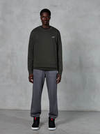 Alpinestars Essentials Ageless Crew, sweat-shirt à col rond vert foncé avec un petit logo blanc sur la poitrine, porté par un mannequin avec un pantalon de survêtement gris et des baskets montantes noires, vêtements lifestyle décontractés