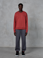 Alpinestars Essentials Ageless Crew, sweat-shirt à col rond rouge brique, porté avec un pantalon de jogging gris et des baskets noires, avec un petit logo noir sur la poitrine, streetwear décontracté d'inspiration sport mécanique