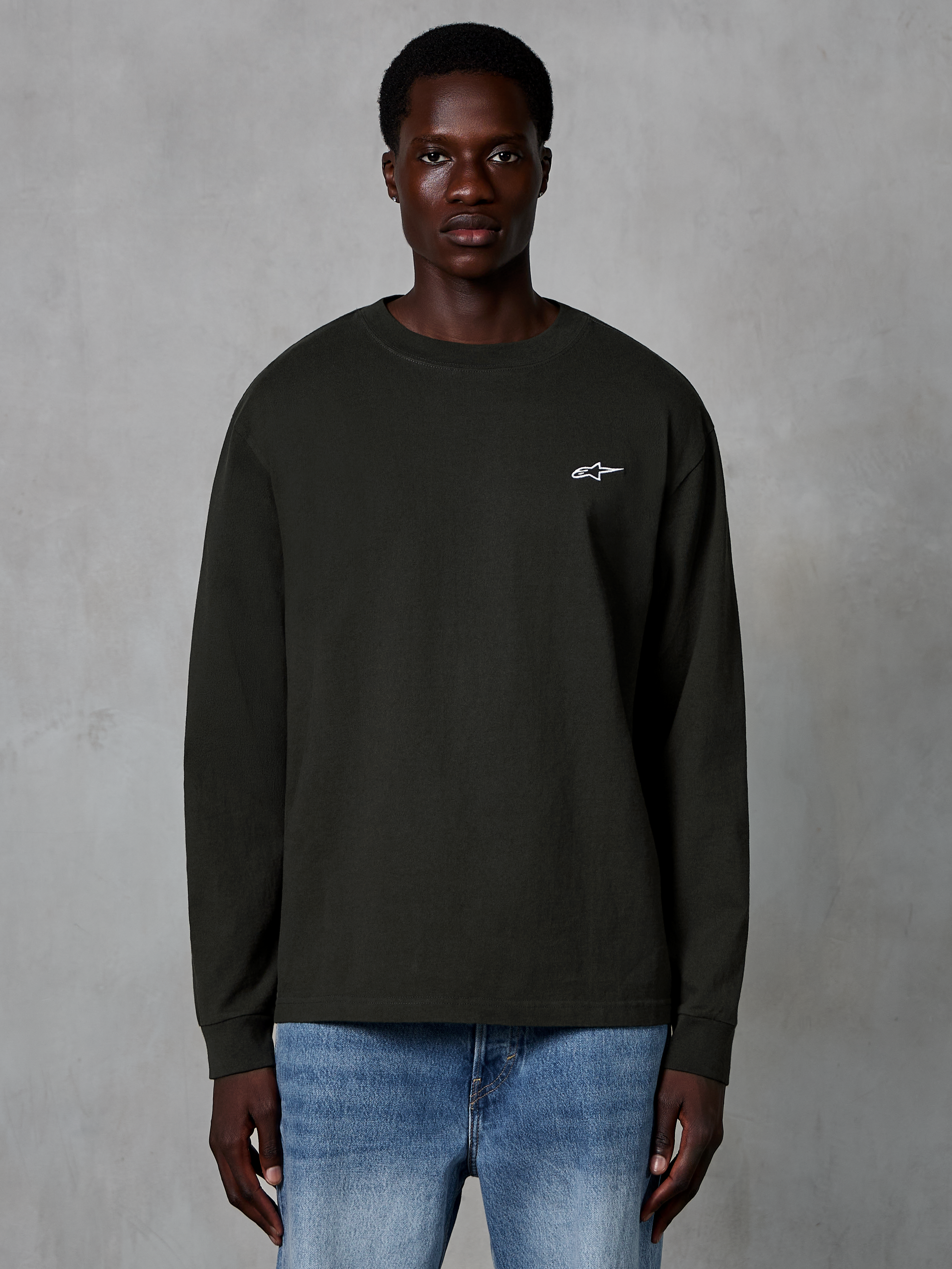 Alpinestars Essentials Ageless LS Tee, t-shirt à manches longues, noir, avec un petit logo Alpinestars blanc sur la poitrine, porté par un mannequin avec un jean bleu, streetwear décontracté en coton biologique