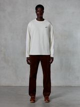 Alpinestars Essentials Ageless LS Tee, Long-Sleeve T-Shirt, Off-White, worn by model with brown corduroy pants and brown leather boots, featuring a small black logo on the chest, relaxed contemporary fit, casual lifestyle streetwear