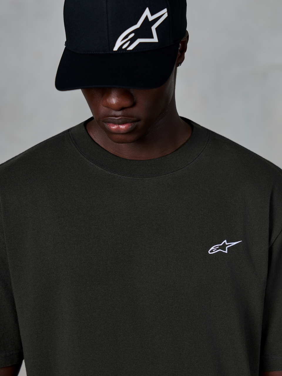 T-shirt Alpinestars Essentials Ageless SS, t-shirt à manches courtes, noir délavé, avec un petit logo Alpinestars blanc sur la poitrine, porté avec une casquette de baseball noire à logo, style streetwear motorsport décontracté