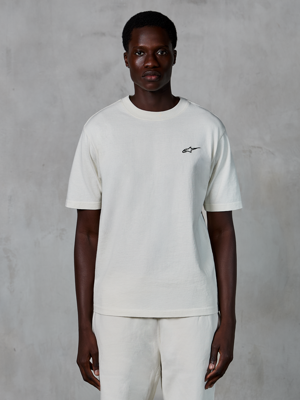 Alpinestars Essentials Ageless SS Tee, t-shirt à manches courtes, blanc cassé, porté par un mannequin homme, doté d'un petit logo Alpinestars noir sur la poitrine, coupe décontractée, jersey de coton biologique, style streetwear motorsport décontracté