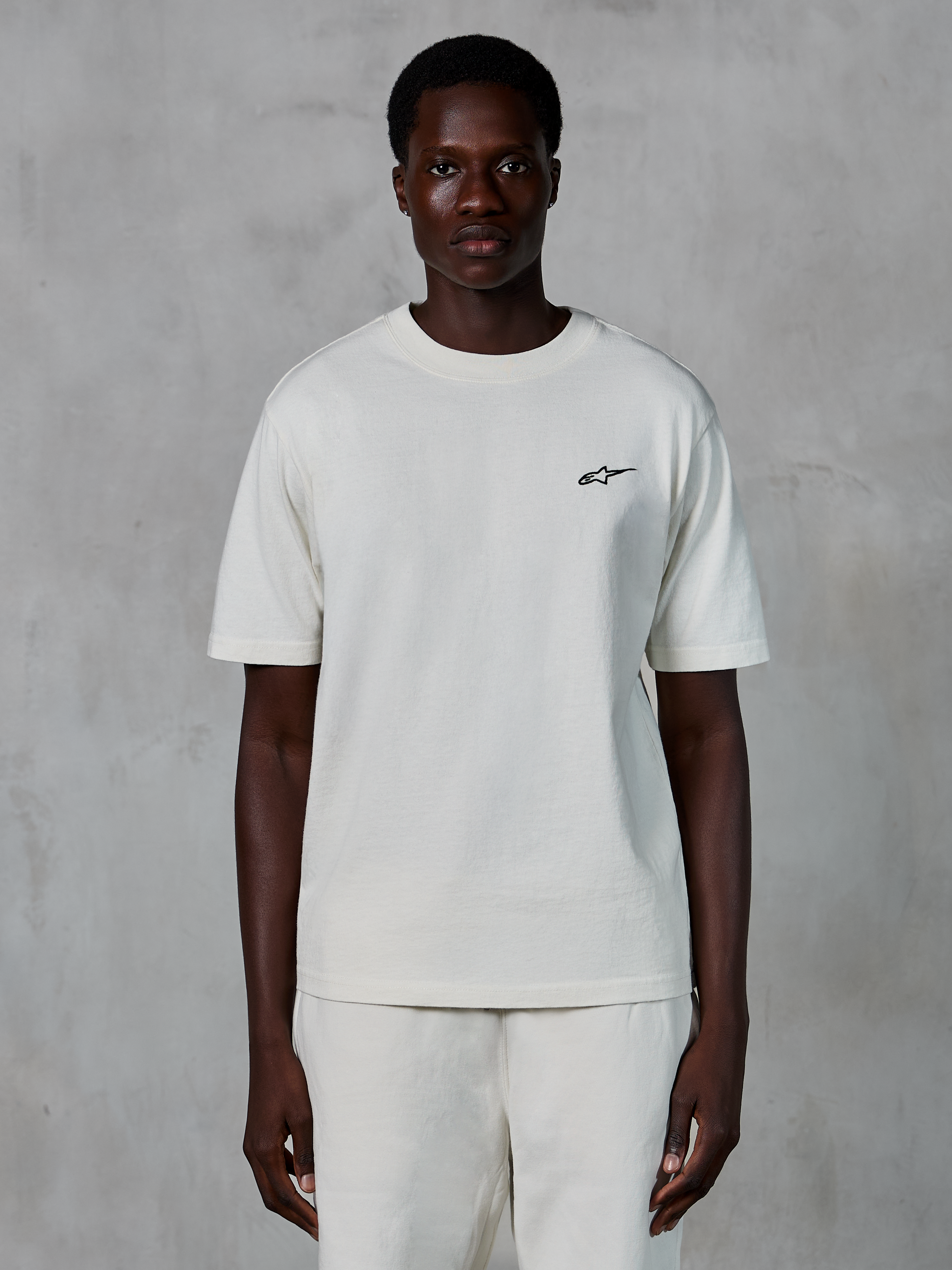 Alpinestars Essentials Ageless SS Tee, t-shirt à manches courtes, blanc cassé, porté par un mannequin homme, doté d'un petit logo Alpinestars noir sur la poitrine, coupe décontractée, jersey de coton biologique, style streetwear motorsport décontracté