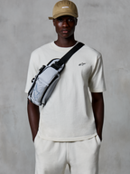 Alpinestars Essentials Ageless SS Tee, t-shirt à manches courtes, blanc cassé, porté avec un short assorti et une casquette beige, arborant un petit logo noir sur la poitrine et un sac bandoulière gris, streetwear décontracté inspiré des sports mécaniques