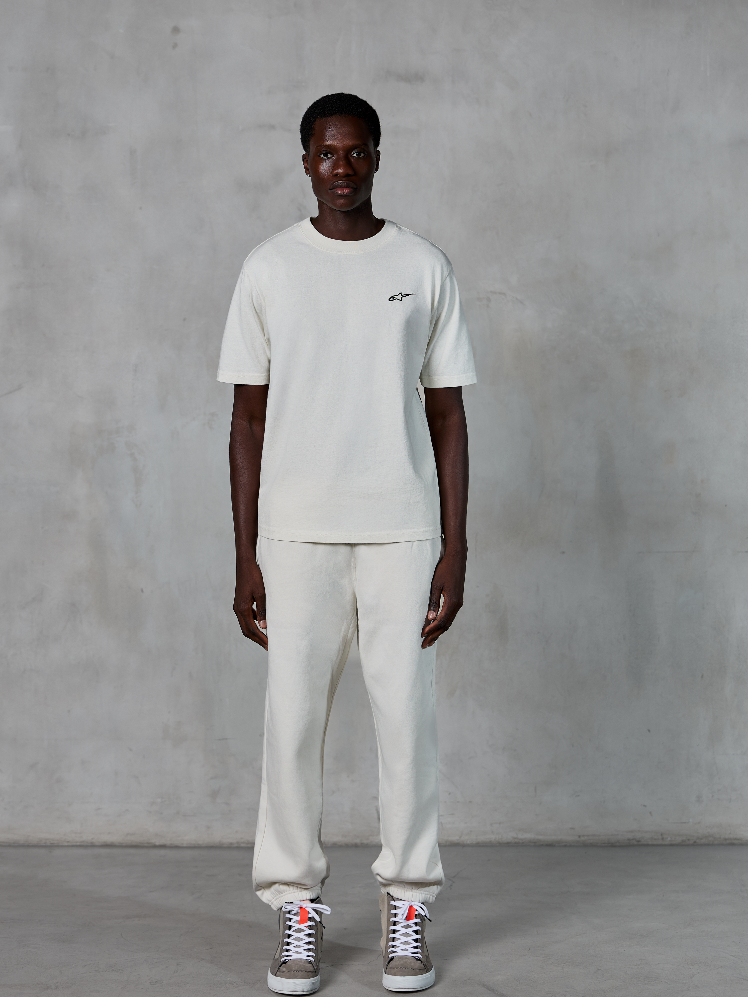 T-shirt Alpinestars Essentials Ageless SS, t-shirt à manches courtes, blanc cassé, arborant un petit logo Alpinestars noir sur la poitrine, porté avec un pantalon de jogging et des baskets assortis pour un look décontracté
