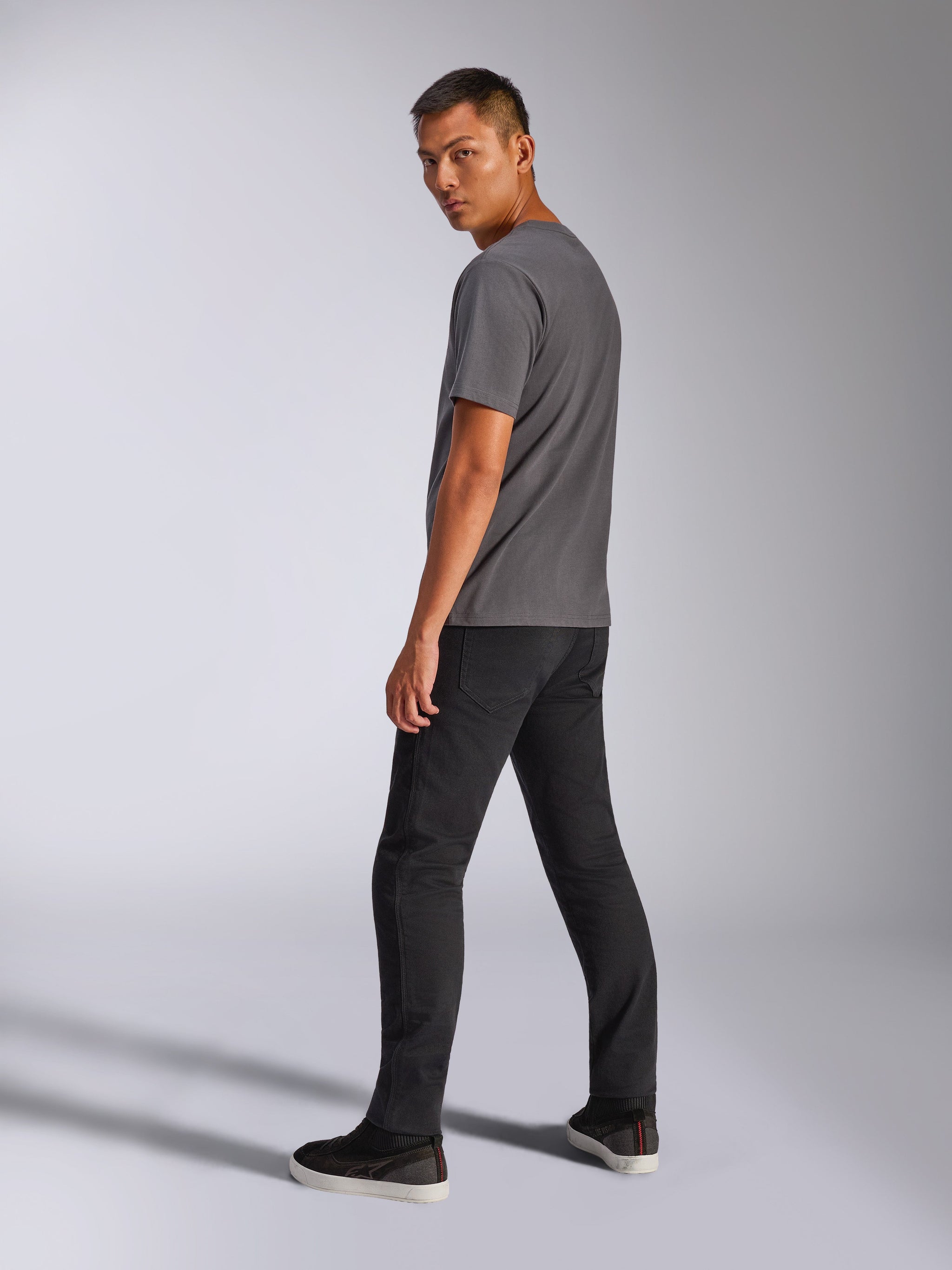 Male model wearing Alpinestars Shaded CSF Tee, Short Sleeve T-Shirt, Charcoal Gray, back view showing the graphic, paired with black pants and sneakers, casual motorsport-inspired streetwear