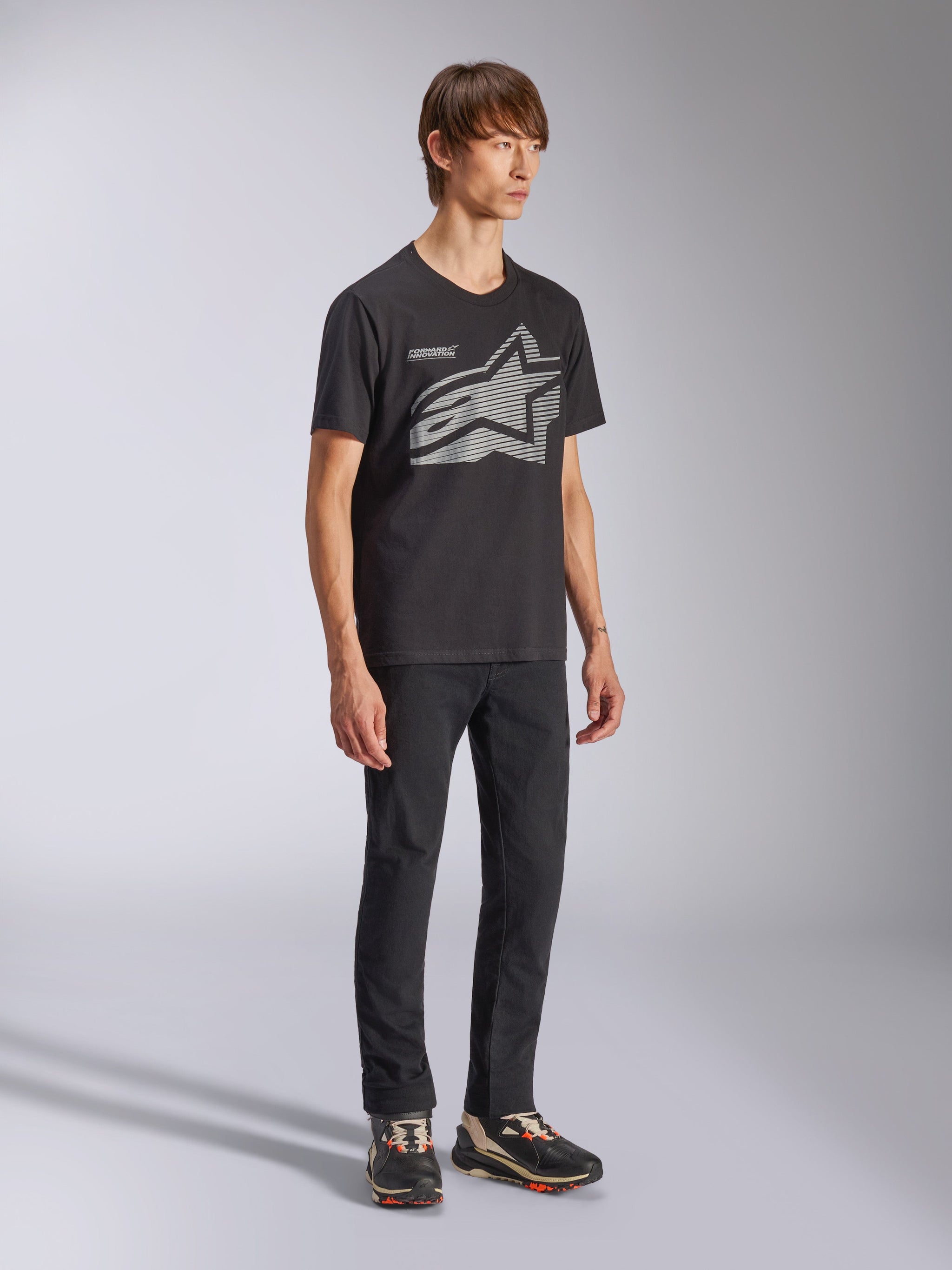 Modèle masculin portant le t-shirt Alpinestars Fasting CSF, t-shirt à manches courtes, noir, logo étoile géométrique Astars gris à l'avant avec texte Forward Innovation, associé à un pantalon noir et des baskets, style sportswear décontracté