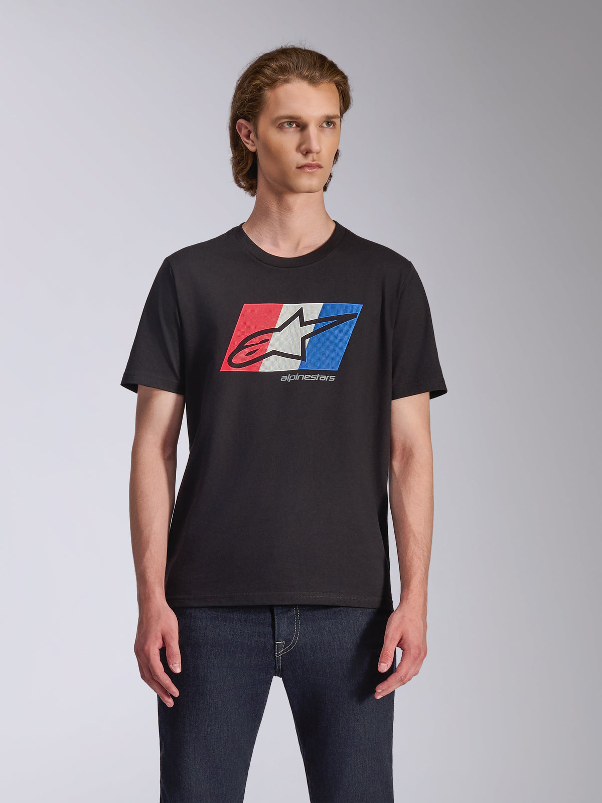 T-shirt Alpinestars Global CSF, t-shirt à manches courtes, noir, graphique logo Ageless rouge, blanc et bleu, porté par un mannequin homme avec un jean foncé, streetwear décontracté motorsport