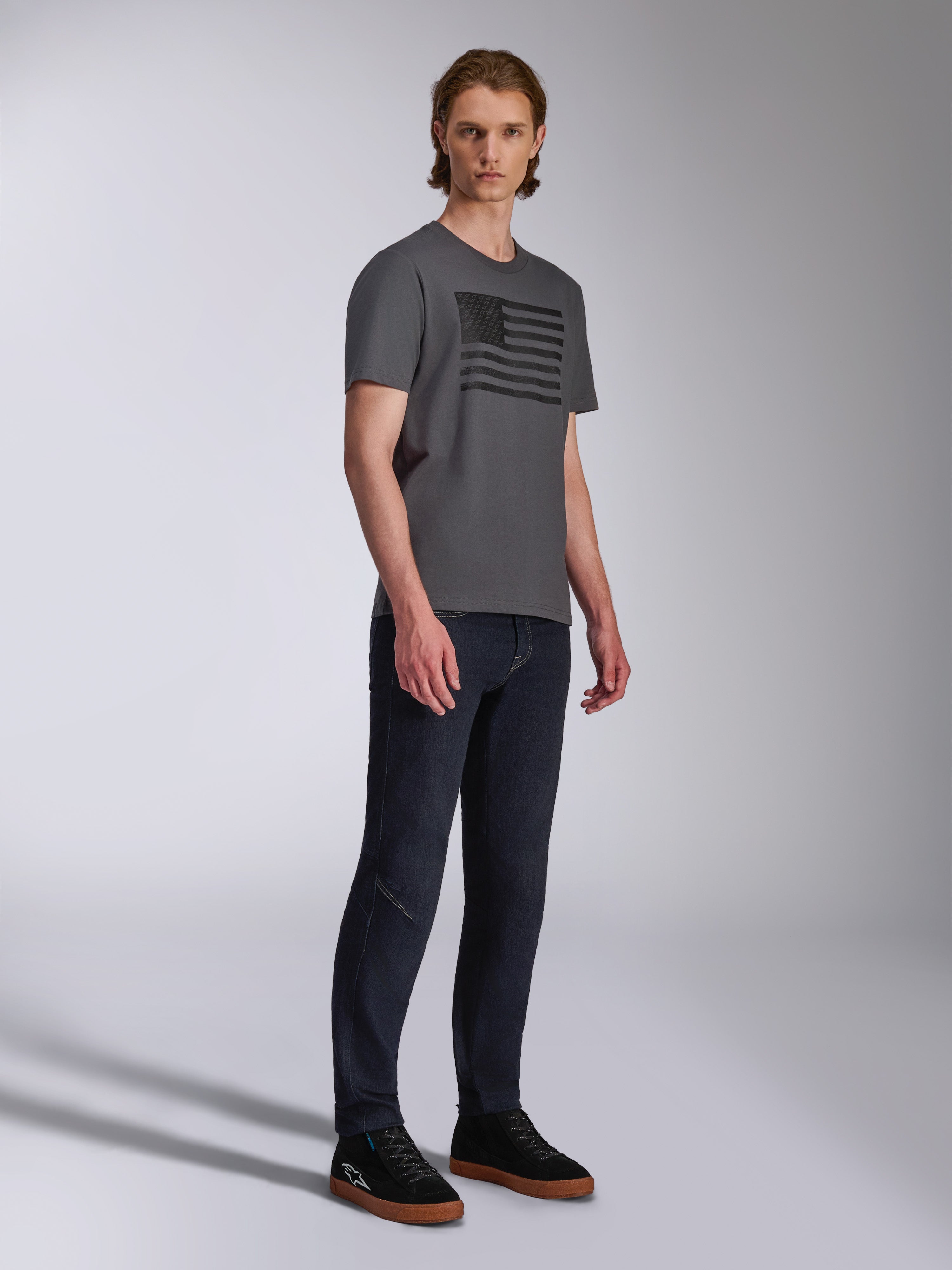 Alpinestars Waving CSF Tee, Short Sleeve T-Shirt, Charcoal, Black Waving Flag Graphic, worn by a model with dark jeans and sneakers, Casual Motorsport Streetwear