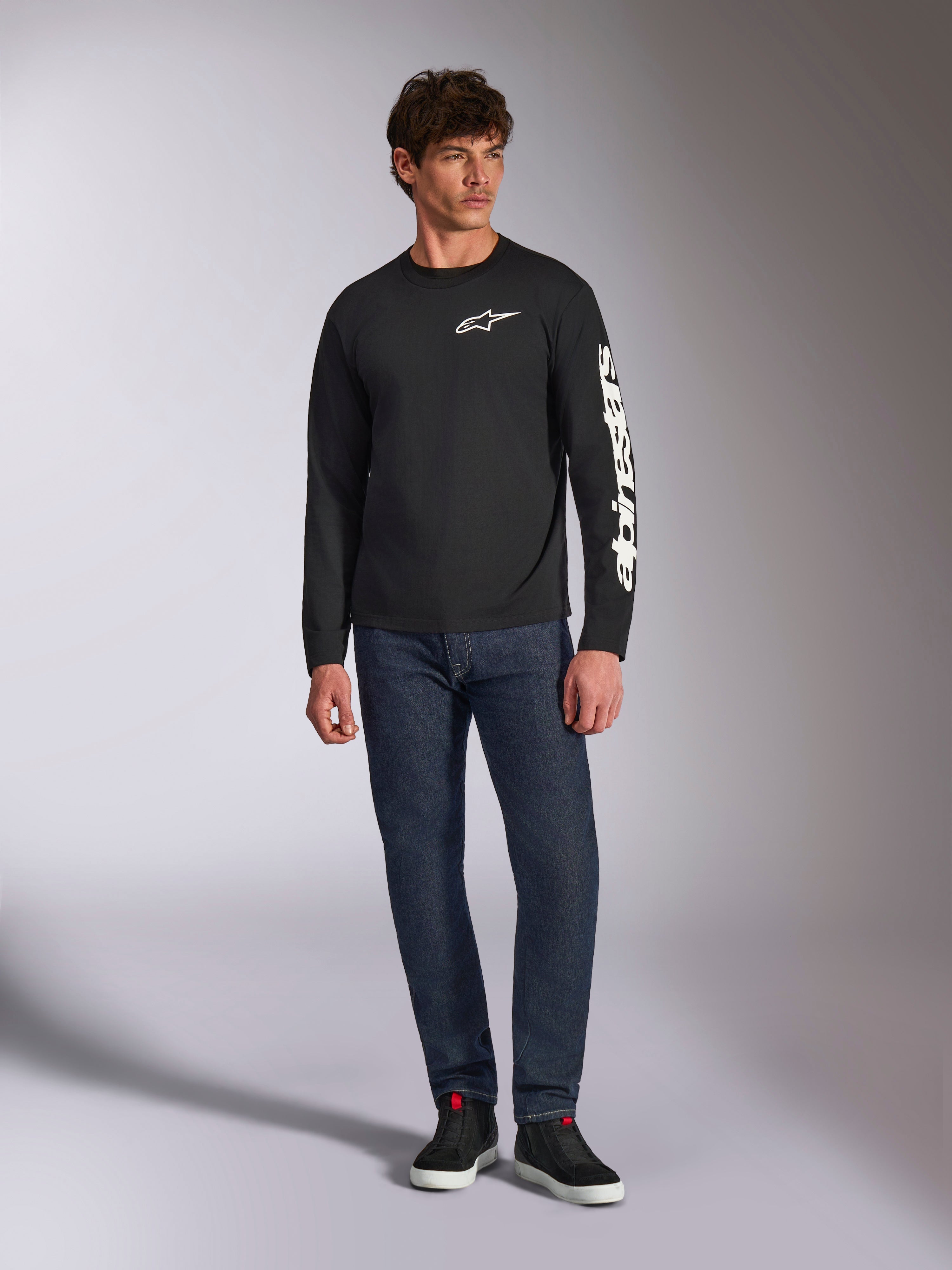 Alpinestars Trackside CSF Tee, t-shirt à manches longues, noir et blanc, porté avec un jean bleu foncé et des baskets noires, arborant le logo Ride 4.0 blanc sur la poitrine et le lettrage Alpinestars sur la manche, streetwear décontracté inspiré des sports mécaniques