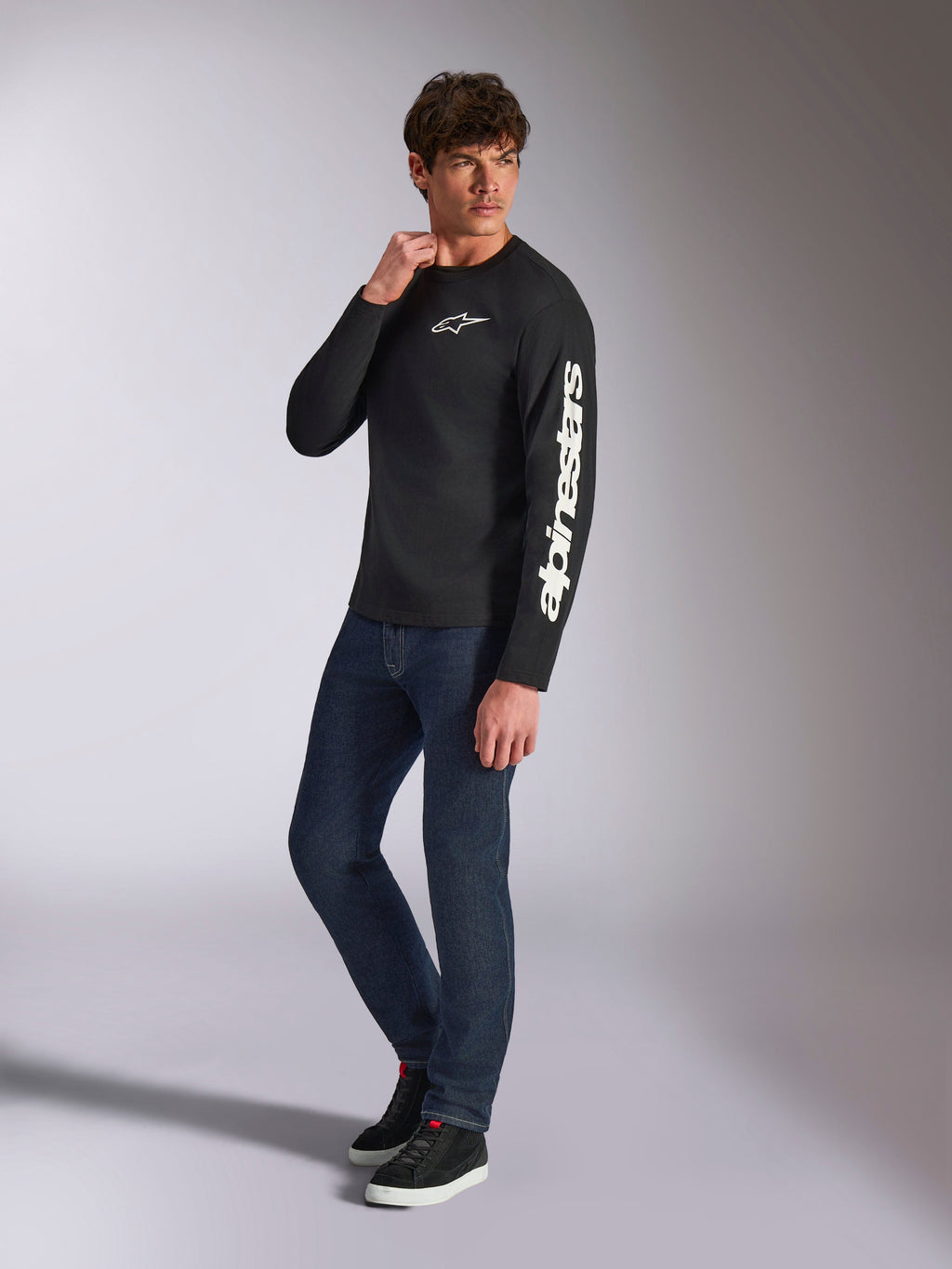 T-shirt à manches longues Alpinestars Trackside CSF, noir, marquage Alpinestars vertical blanc sur la manche et logo sur la poitrine, porté par un mannequin homme avec un jean bleu foncé et des baskets, style streetwear décontracté inspiré du sport automobile