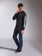 T-shirt à manches longues Alpinestars Trackside CSF, noir, marquage Alpinestars vertical blanc sur la manche et logo sur la poitrine, porté par un mannequin homme avec un jean bleu foncé et des baskets, style streetwear décontracté inspiré du sport automobile