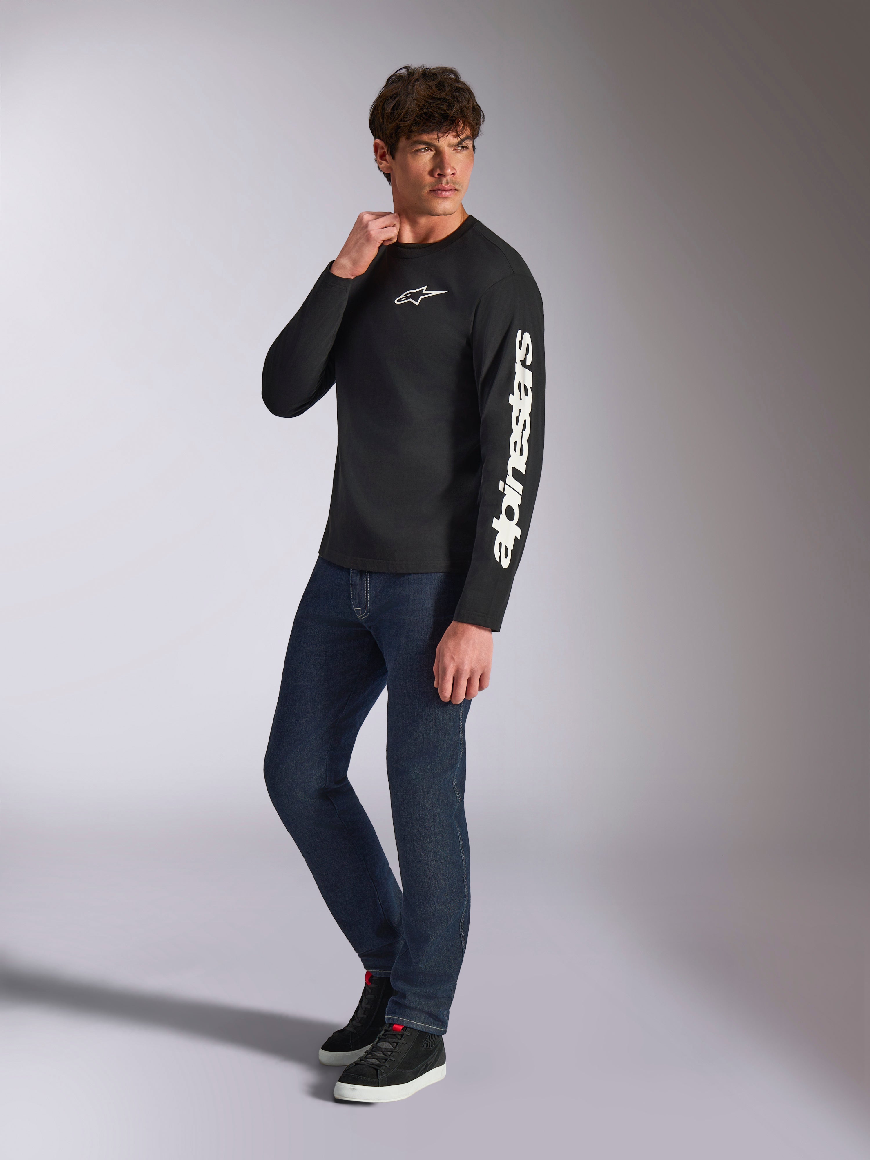 T-shirt à manches longues Alpinestars Trackside CSF, noir, marquage Alpinestars vertical blanc sur la manche et logo sur la poitrine, porté par un mannequin homme avec un jean bleu foncé et des baskets, style streetwear décontracté inspiré du sport automobile