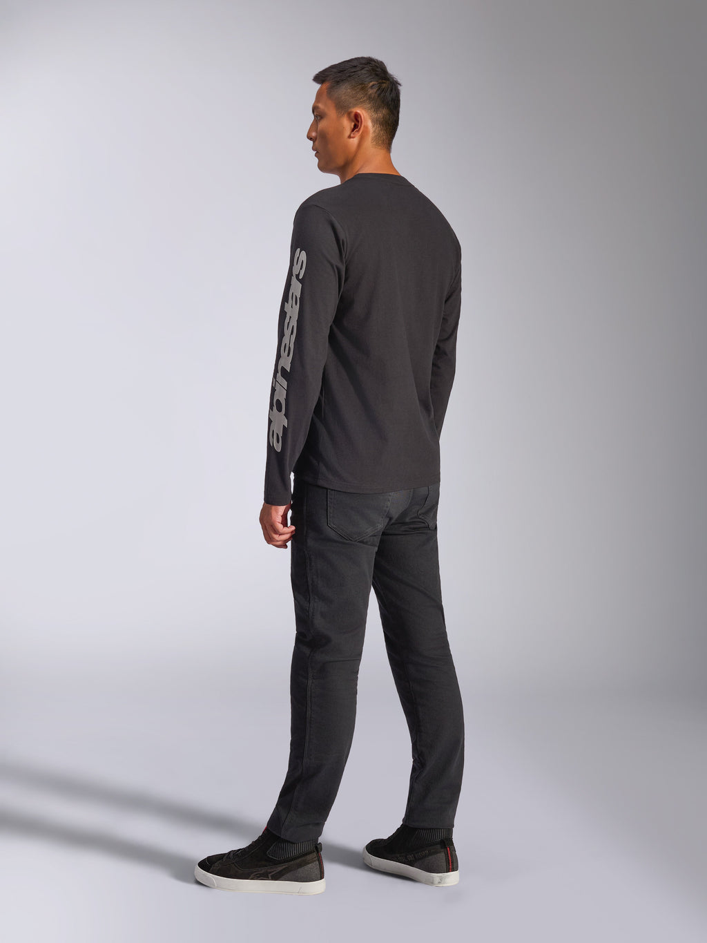 T-shirt Alpinestars Trackside CSF à manches longues, noir, avec un grand logo Alpinestars gris sur la manche gauche, porté par un mannequin avec un pantalon noir et des baskets décontractées, streetwear décontracté inspiré du sport automobile
