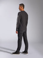 T-shirt Alpinestars Trackside CSF à manches longues, noir, avec un grand logo Alpinestars gris sur la manche gauche, porté par un mannequin avec un pantalon noir et des baskets décontractées, streetwear décontracté inspiré du sport automobile