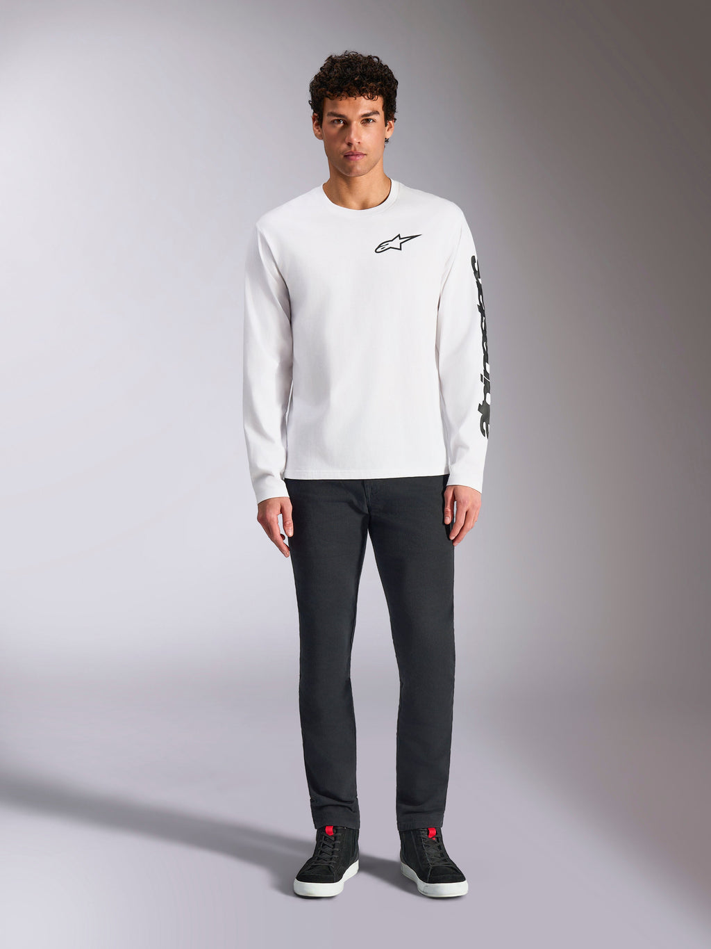 T-shirt à manches longues Alpinestars Trackside CSF, blanc avec logos noirs, petit logo sur la poitrine et texte vertical sur la manche, porté par un mannequin avec un pantalon sombre et des baskets noires, streetwear décontracté inspiré du sport automobile