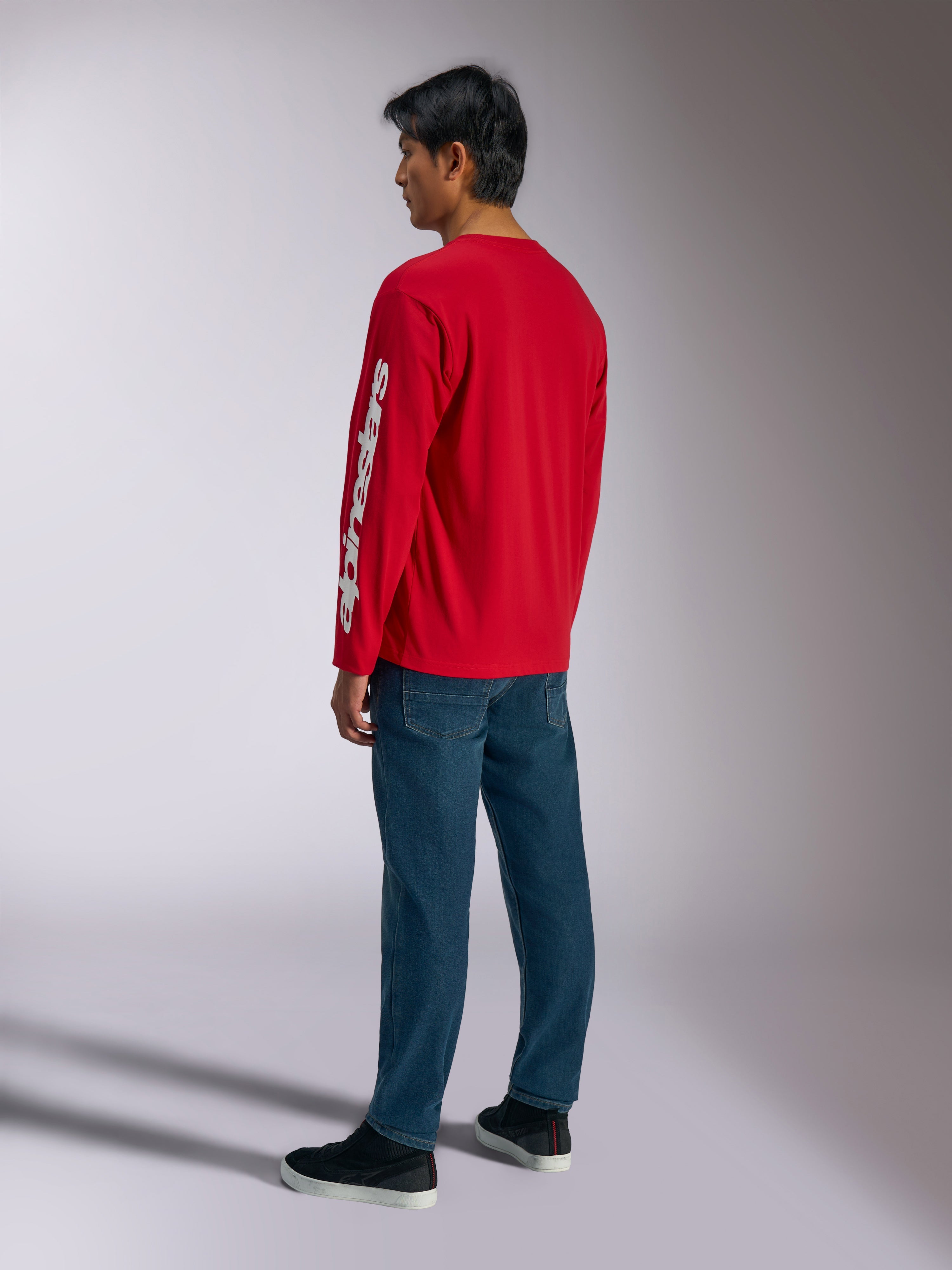 T-shirt à manches longues Alpinestars Trackside CSF, rouge, arborant un logo Alpinestars vertical blanc sur la manche, porté par un mannequin avec un jean bleu et des baskets noires, style streetwear décontracté inspiré par les sports mécaniques