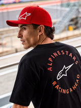 Casquette Alpinestars Corp Shift Multi, casquette rouge à visière courbée avec logo Astars 3D blanc, portée avec un t-shirt Alpinestars noir arborant le graphisme « Racing Ahead », cadre lifestyle motorsport sur un circuit de course