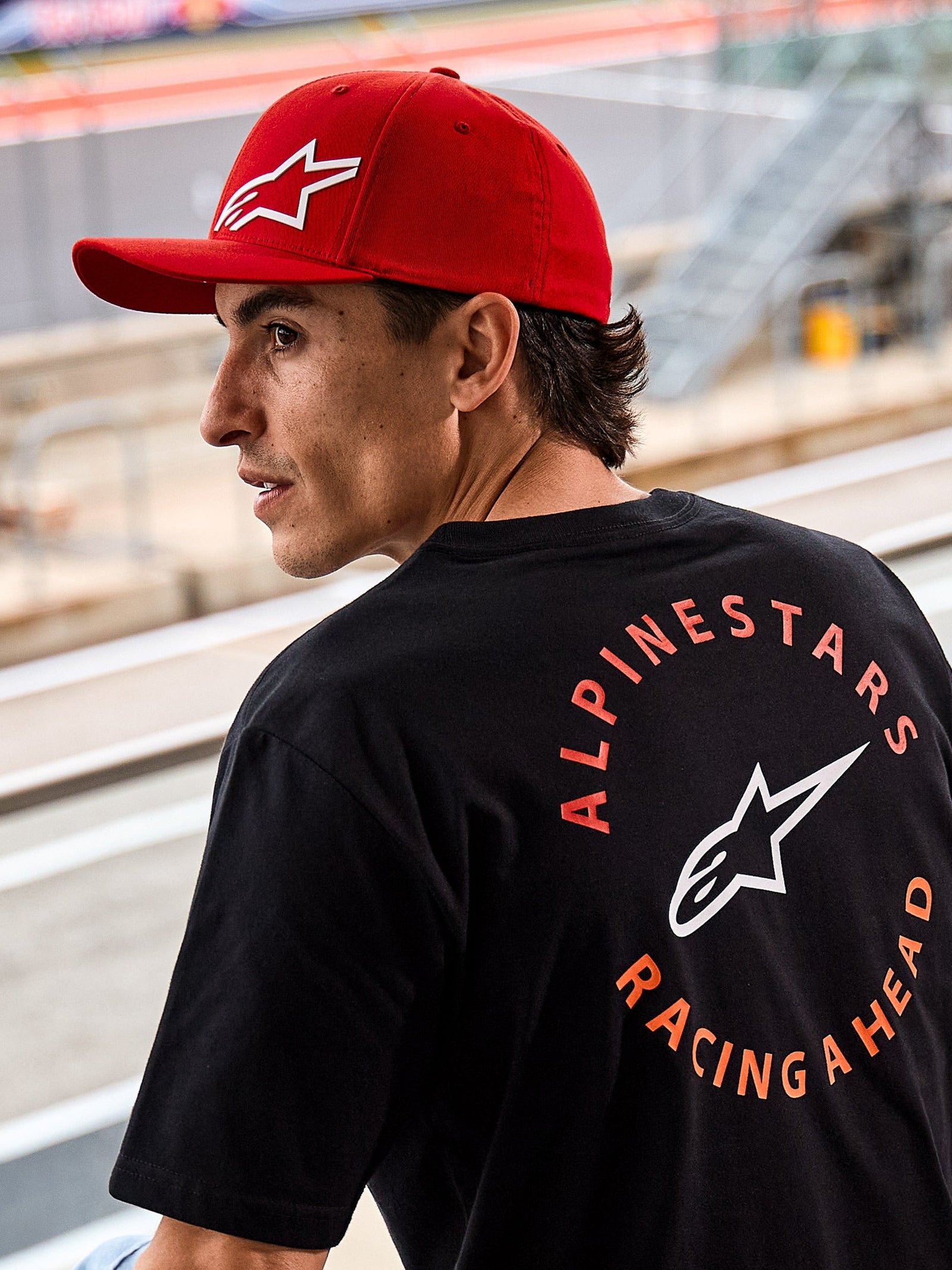 Casquette Alpinestars Corp Shift Multi, casquette rouge à visière courbée avec logo Astars 3D blanc, portée avec un t-shirt Alpinestars noir arborant le graphisme « Racing Ahead », cadre lifestyle motorsport sur un circuit de course