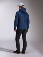 Sweat à capuche Alpinestars Frontal, vue arrière d'un sweat à capuche en polaire bleu marine, logo en lettres blanc sur la manche, porté avec un pantalon noir et une casquette trucker blanche en maille filet, streetwear décontracté inspiré du sport automobile