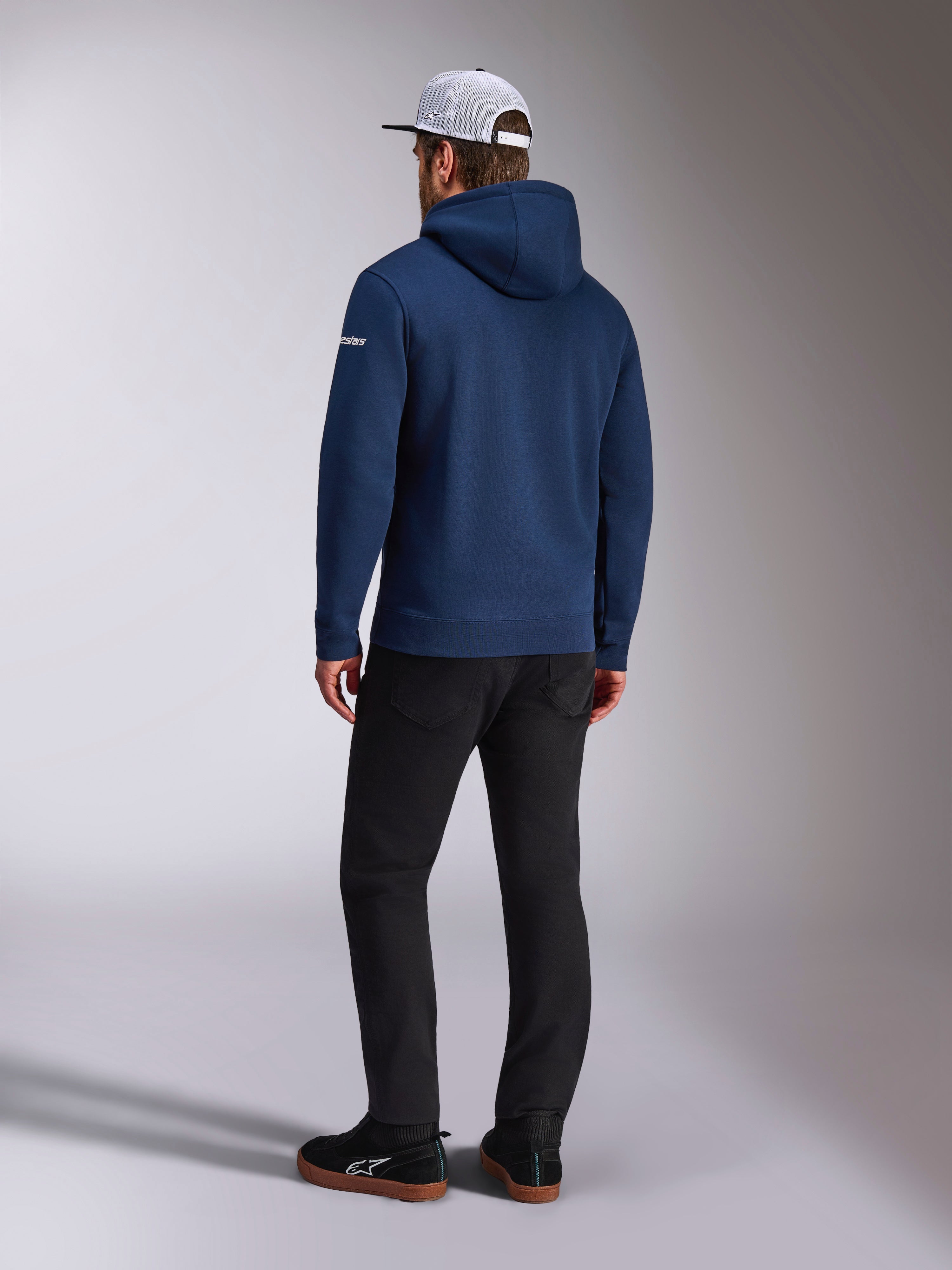 Sweat à capuche Alpinestars Frontal, vue arrière d'un sweat à capuche en polaire bleu marine, logo en lettres blanc sur la manche, porté avec un pantalon noir et une casquette trucker blanche en maille filet, streetwear décontracté inspiré du sport automobile