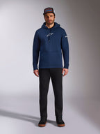 Modèle masculin portant le sweat à capuche Frontal Alpinestars bleu marine, polaire à enfiler avec logo Astars blanc sur la poitrine et logo lettrage sur la manche, doté d'une poche kangourou et d'une capuche à cordon, associé à un pantalon noir et une casquette de marque, streetwear motorsport décontracté