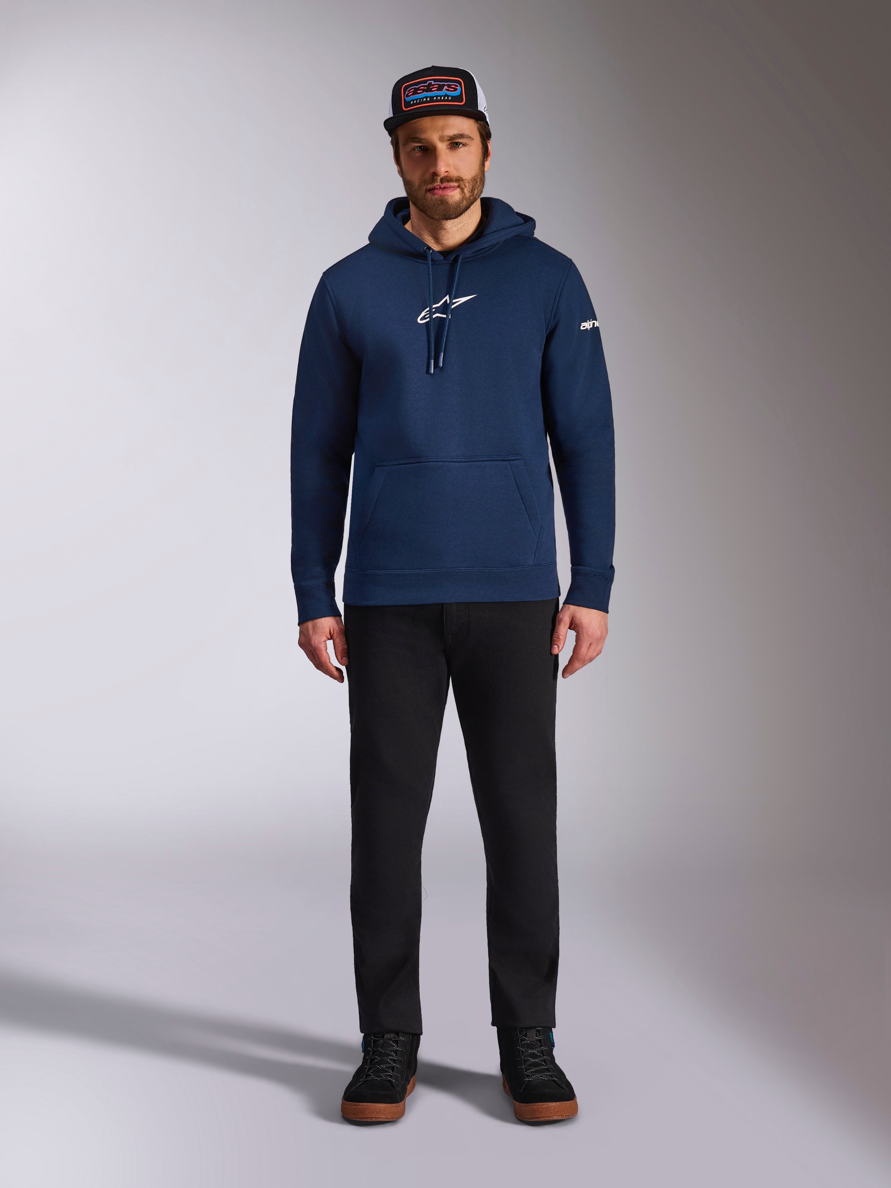Modèle masculin portant le sweat à capuche Frontal Alpinestars bleu marine, polaire à enfiler avec logo Astars blanc sur la poitrine et logo lettrage sur la manche, doté d'une poche kangourou et d'une capuche à cordon, associé à un pantalon noir et une casquette de marque, streetwear motorsport décontracté