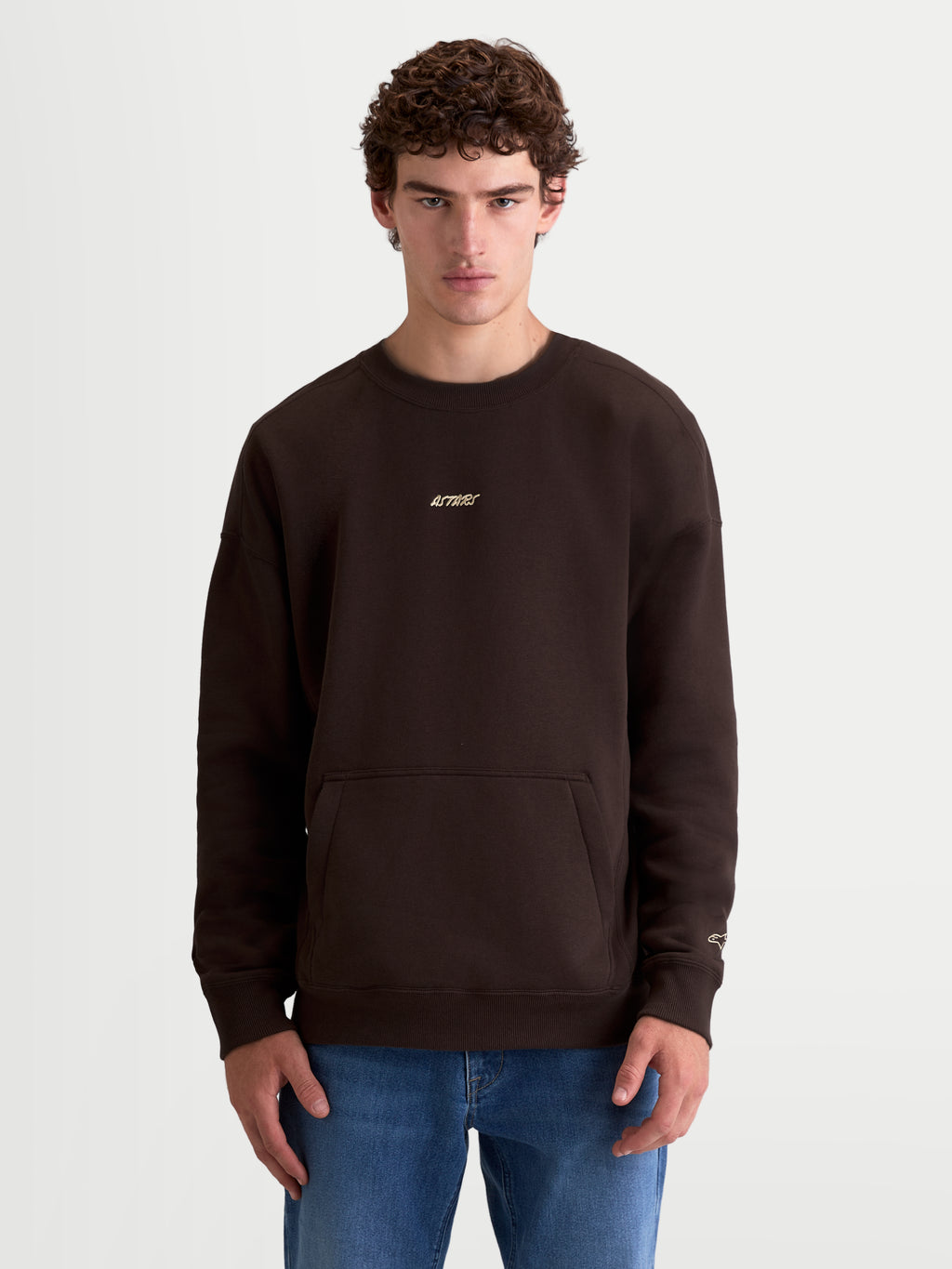 Alpinestars Guise Oversize Sweat, pull en polaire marron de style sportswear, coupe mixte décontractée sur modèle, coton et polyester recyclé, avec poche kangourou et logos Alpinestars brodés sur la poitrine et la manche.
