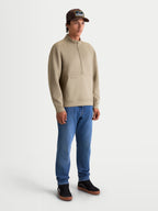 Alpinestars sweatshirt demi-zip Apera, pull sportswear pour homme, beige, modèle portant le vêtement avec un jean bleu, fermeture demi-zippée, poche kangourou et logo ton sur ton, style décontracté inspiré du sport automobile