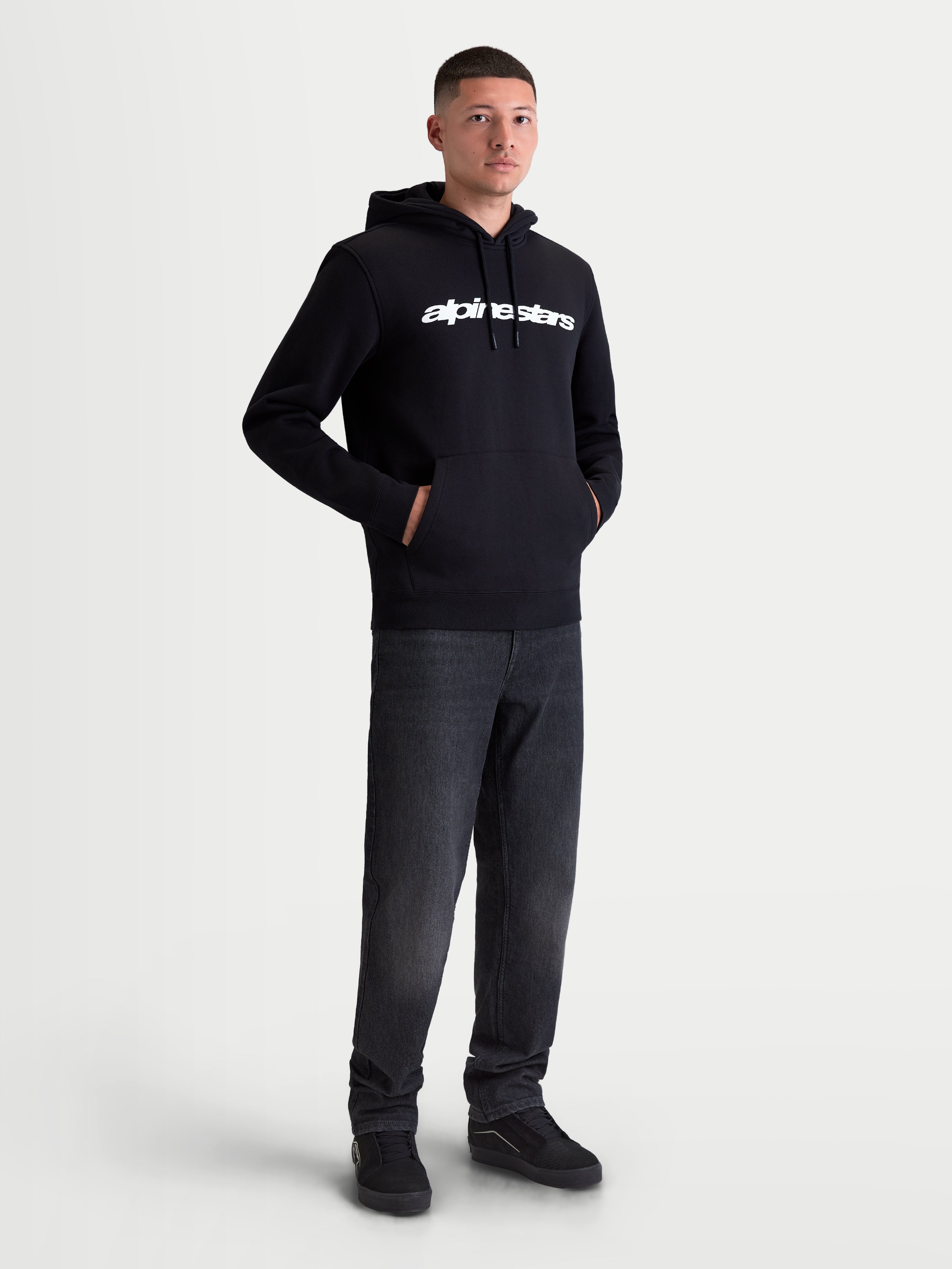Alpinestars Sweat à capuche Race Wordmark, sweat à capuche polaire de sportswear pour homme, noir et blanc, logo linéaire blanc sur la poitrine, capuche avec cordons de serrage et poche kangourou frontale, porté par un modèle.