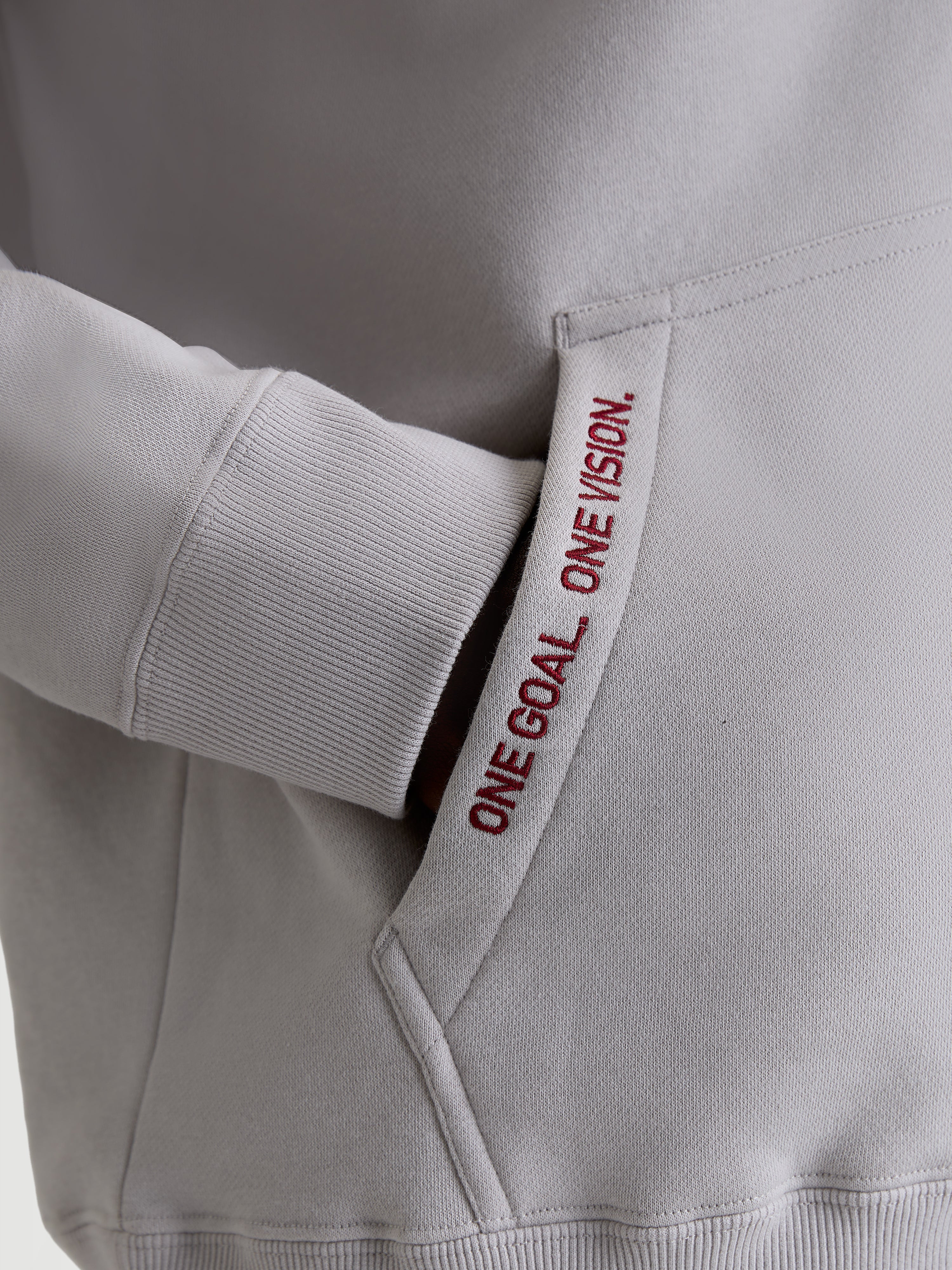 Alpinestars Sweat à capuche Pioneer Oversized, polaire sportswear grise et rouge, détail de la poche kangourou avec texte brodé 'ONE GOAL. ONE VISION.' et poignet côtelé, vêtement lifestyle premium pour passionnés de sports mécaniques