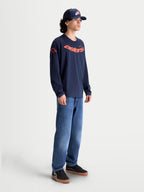 Alpinestars t-shirt Velocifier CSF, t-shirt à manches longues pour homme en bleu marine et orange, mélange de coton et polyester recyclé avec graphisme Astars incurvé et logos sur les épaules, style sportswear décontracté porté par un modèle.