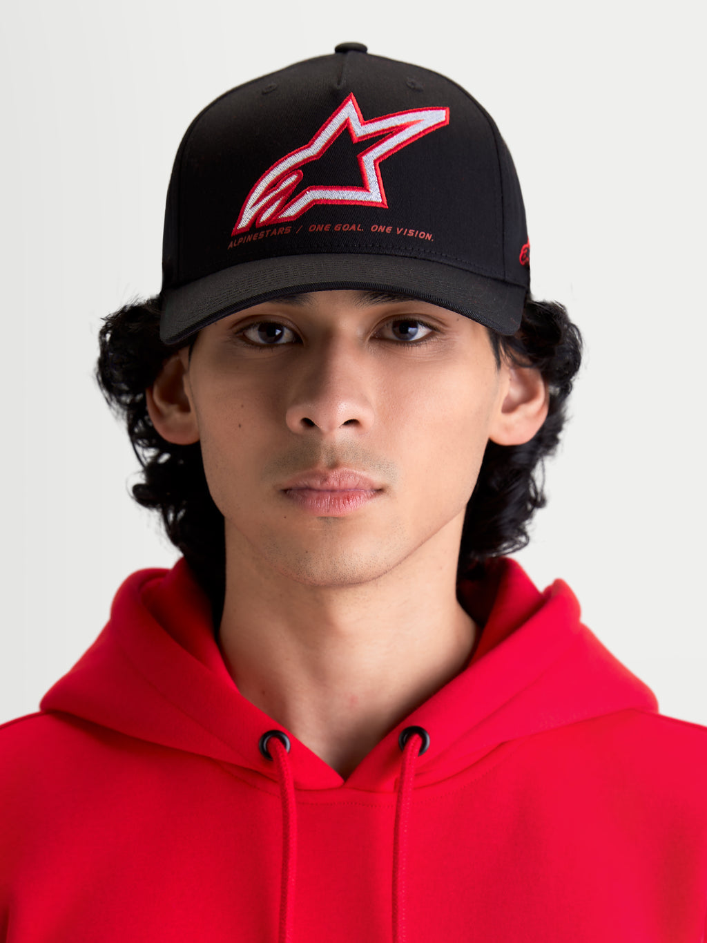 Alpinestars Lineout, casquette de style sportswear, noire avec logo Astars brodé rouge et blanc et slogan One Goal One Vision, couronne structurée et visière incurvée, vue de face sur un modèle portant un sweat à capuche rouge, construction en sergé