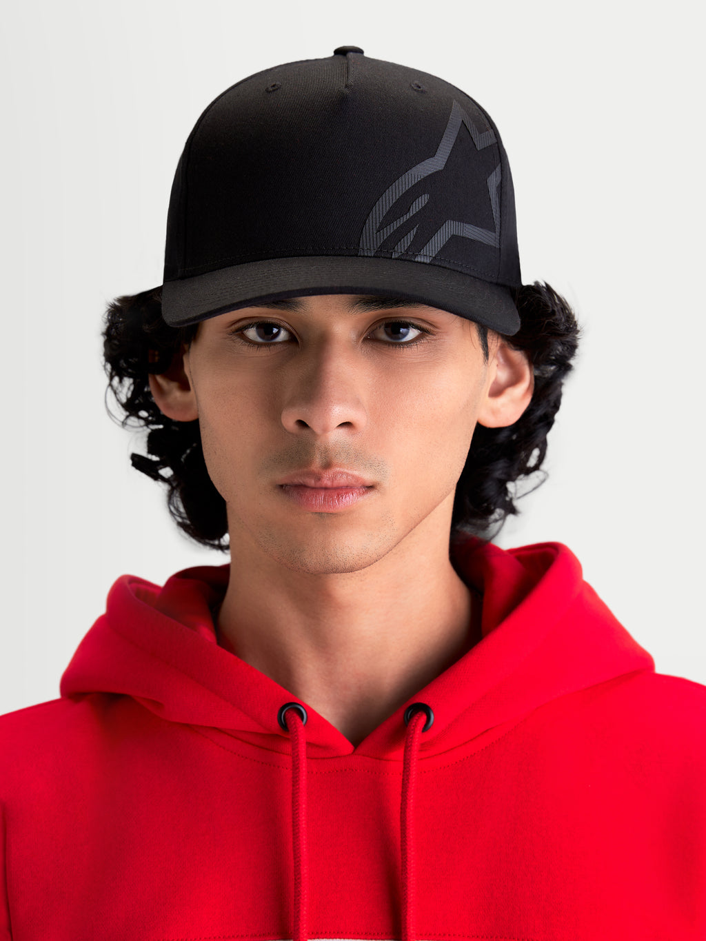 Alpinestars casquette Maze, casquette de sportswear noire inspirée du sport automobile, vue de face sur un modèle portant un sweat à capuche rouge. Dotée d'une couronne structurée, d'une visière incurvée et d'un logo Astars Corp Shift texturé en sergé pour un style décontracté.