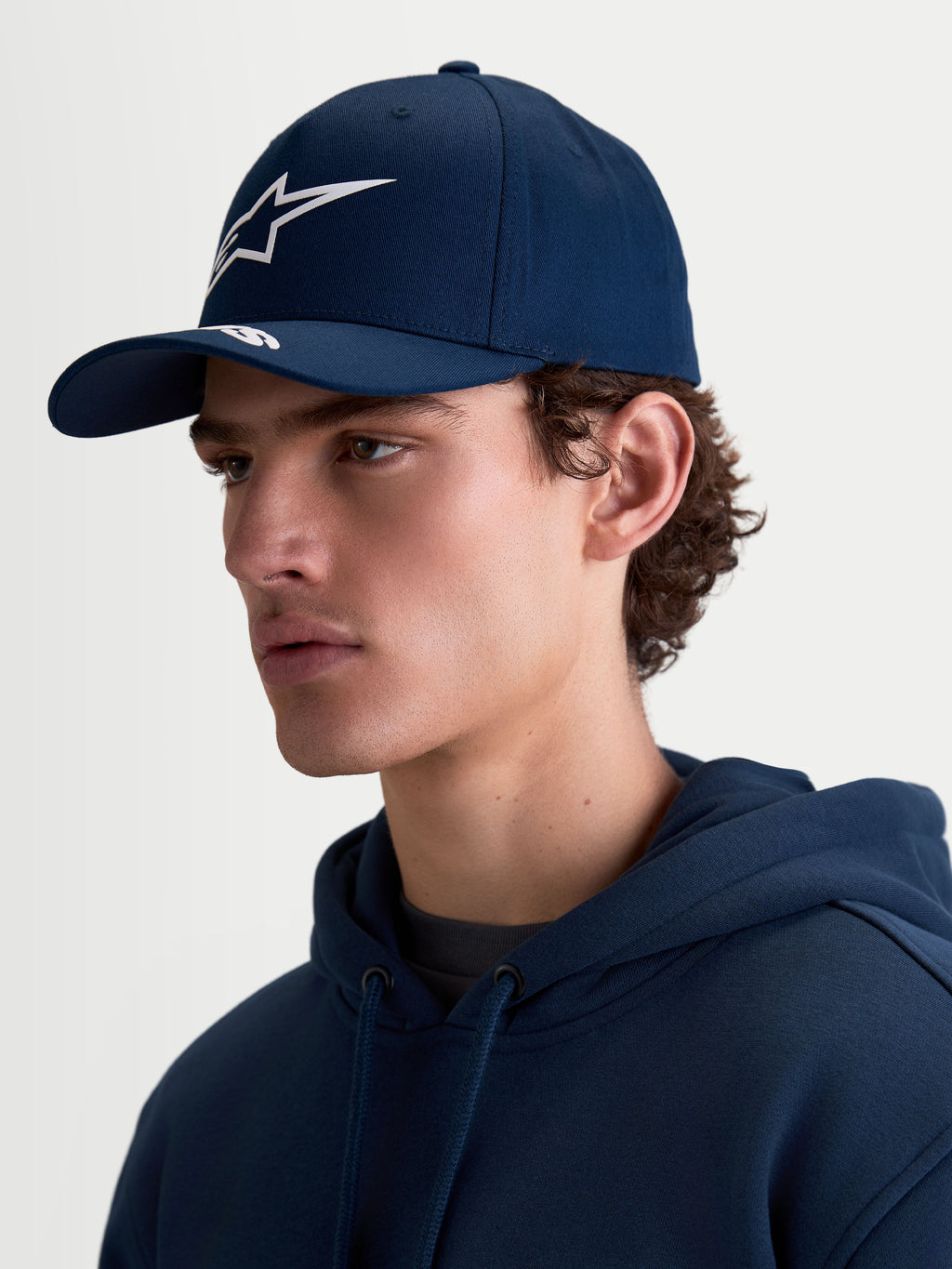 Alpinestars Arclight Hat, Navy Blue Sportswear Baseball Cap for Men and Women, featuring a curved bill with white puff-print Astars branding and a structured crown. On-model side profile view showing the hat's classic motorsport-inspired fit and durable cotton twill fabric.